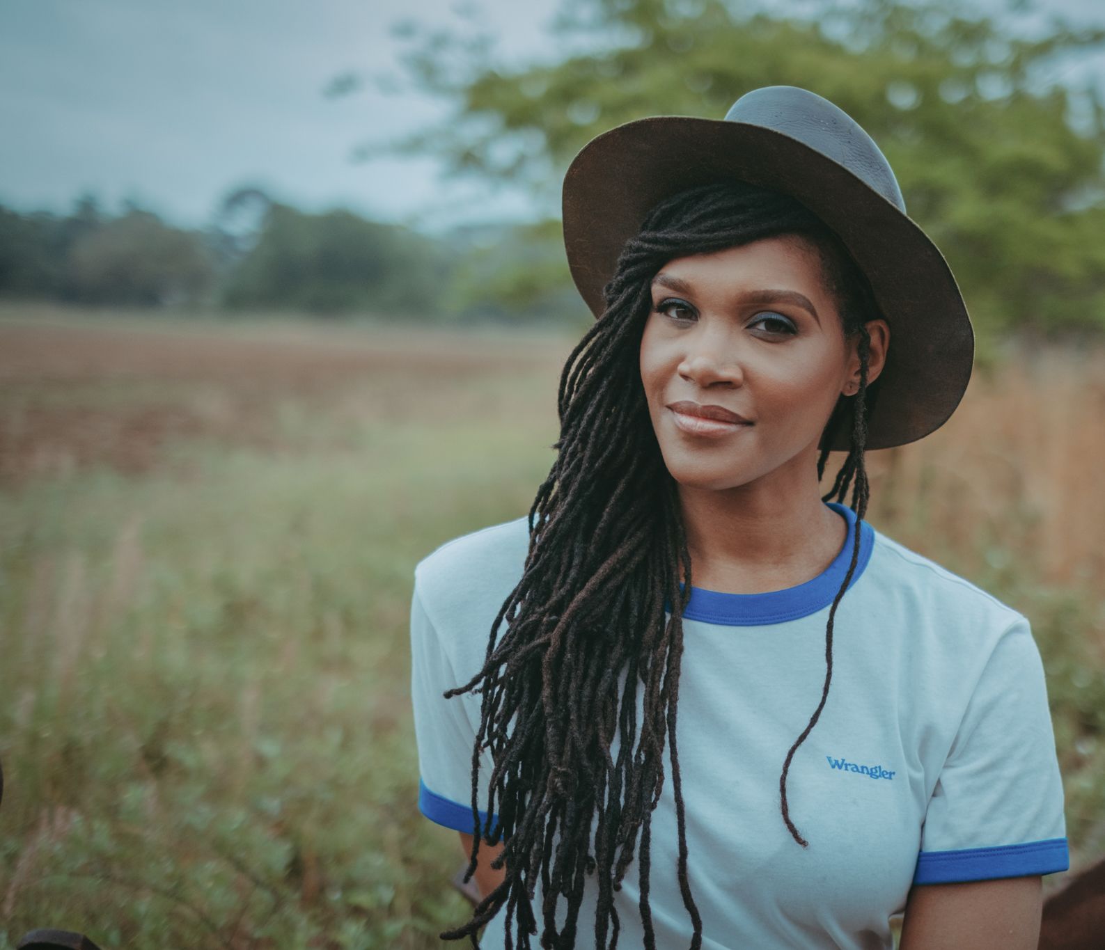 Candace Dantes, wearing a brimmed hat and t-shirt, stands in a field for her professional headshot