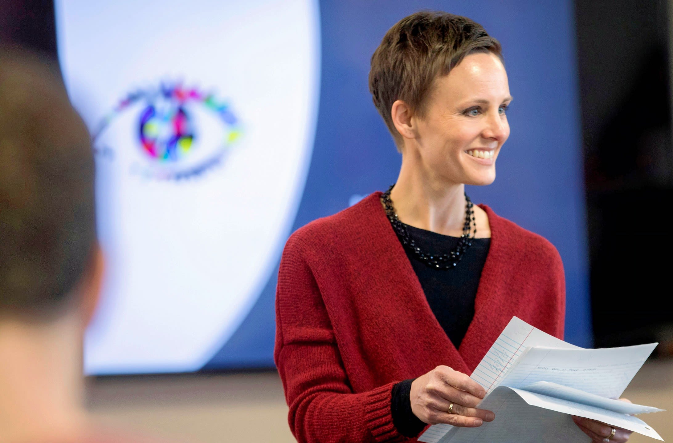 Standing in front of a smart board, smiling and wearing a black shirt with a red sweater, Abigail Borron hands out papers