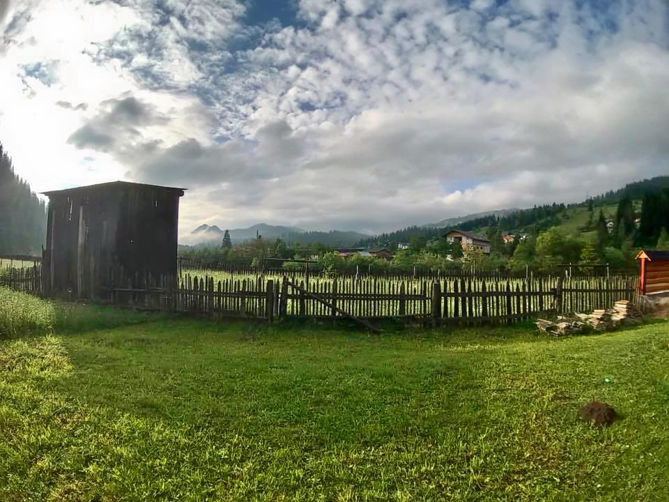 With clouds overhead, a small shed and wooden fence are in the foreground with a green pasture and softly rolling hills in the background.