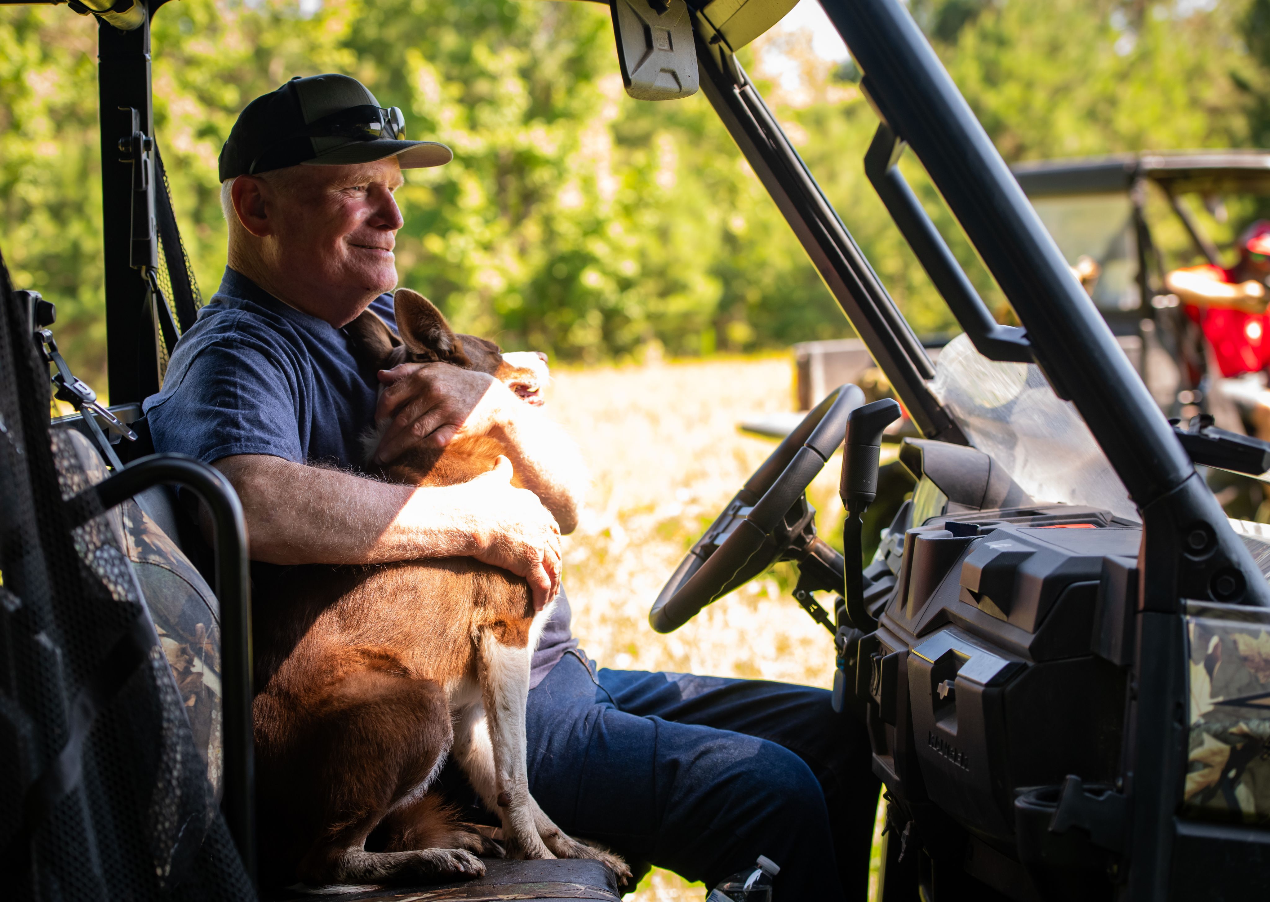 A man sits in the cab of a tractor hugging a brown and white cattle dog.