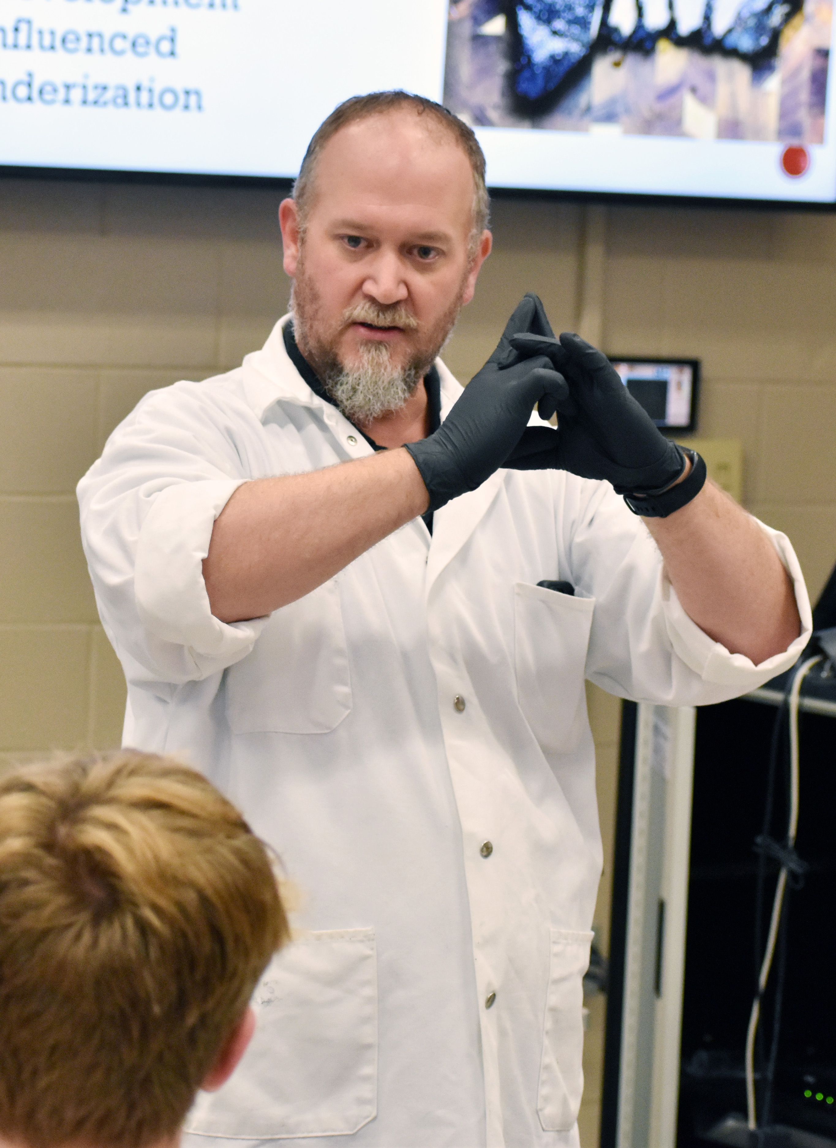 Stelzleni, wearing gloves and standing in front of a projector screen, presents to students.