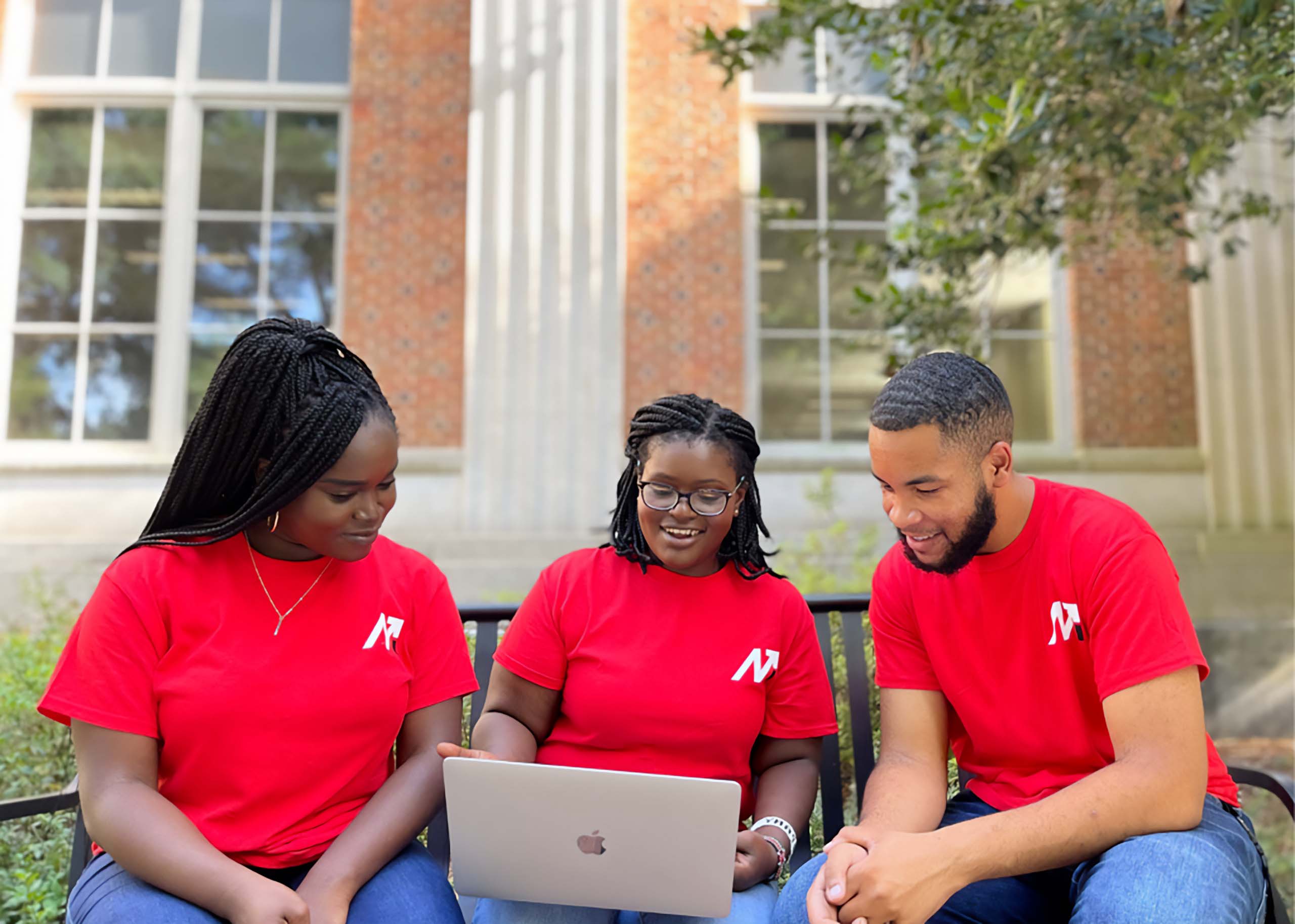 From left, UGA students Ifeoluwa Adu, Tiffany Mukundi and Eric Okanume sit on a UGA campus bench looking at a Mac laptop. All three are wearing red Mentors on a Mission t-shirts.