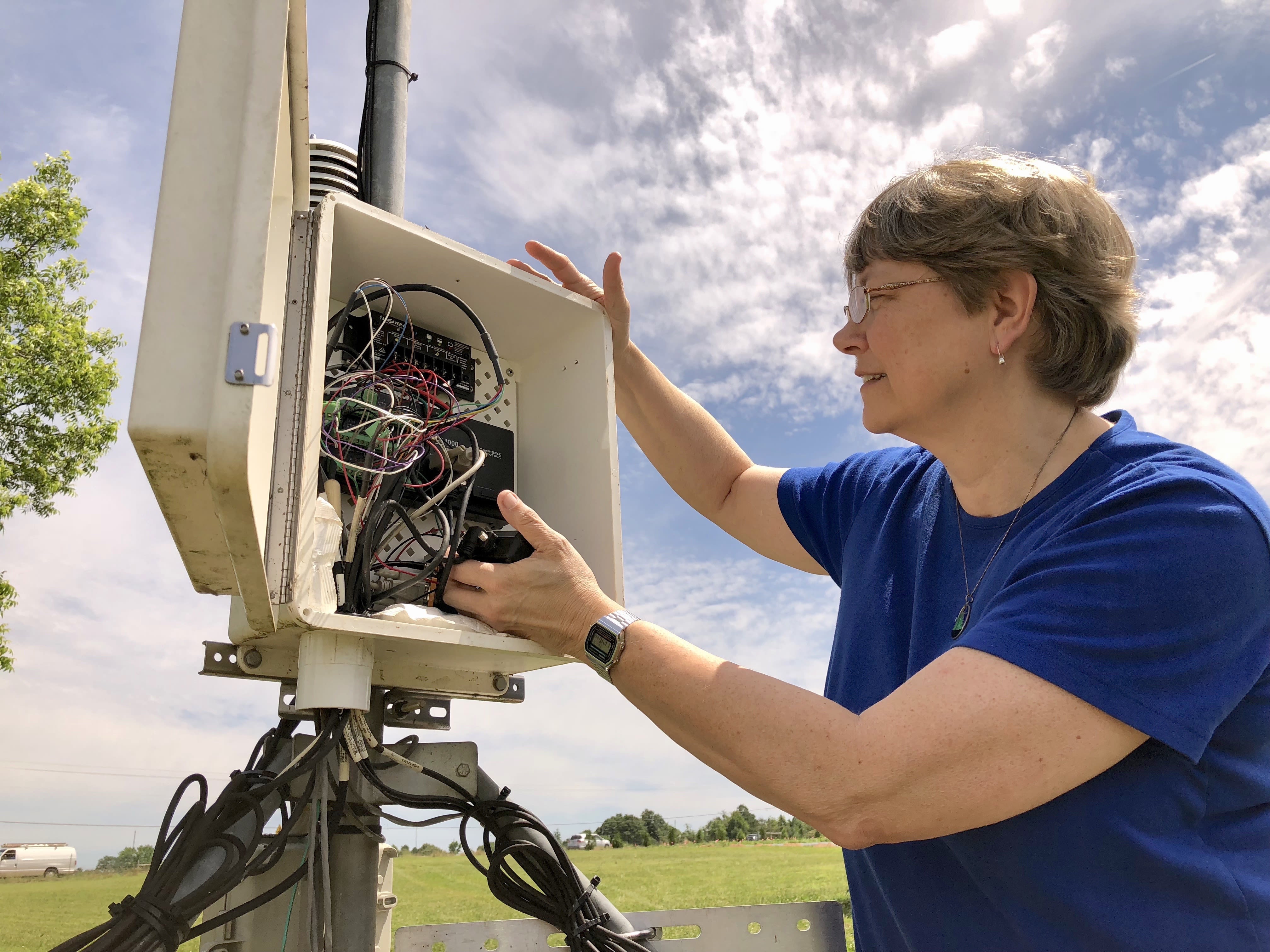 A woman with short hair and glasses, wearing a blue shirt, checks a weather monitoring station.