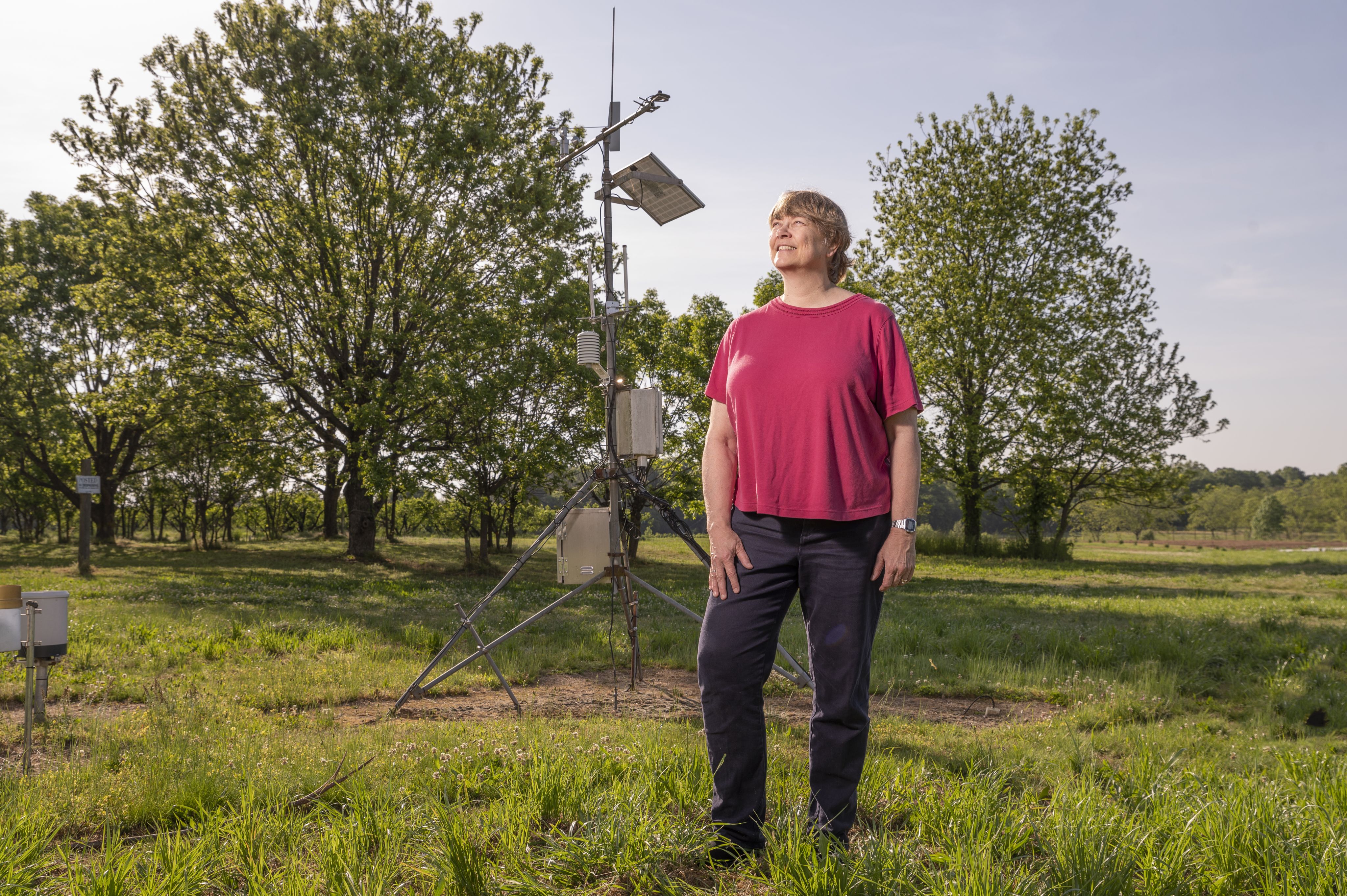 A smiling woman in a red shirt and black plants stands in front of a weather monitoring station in a green field with trees in the background.