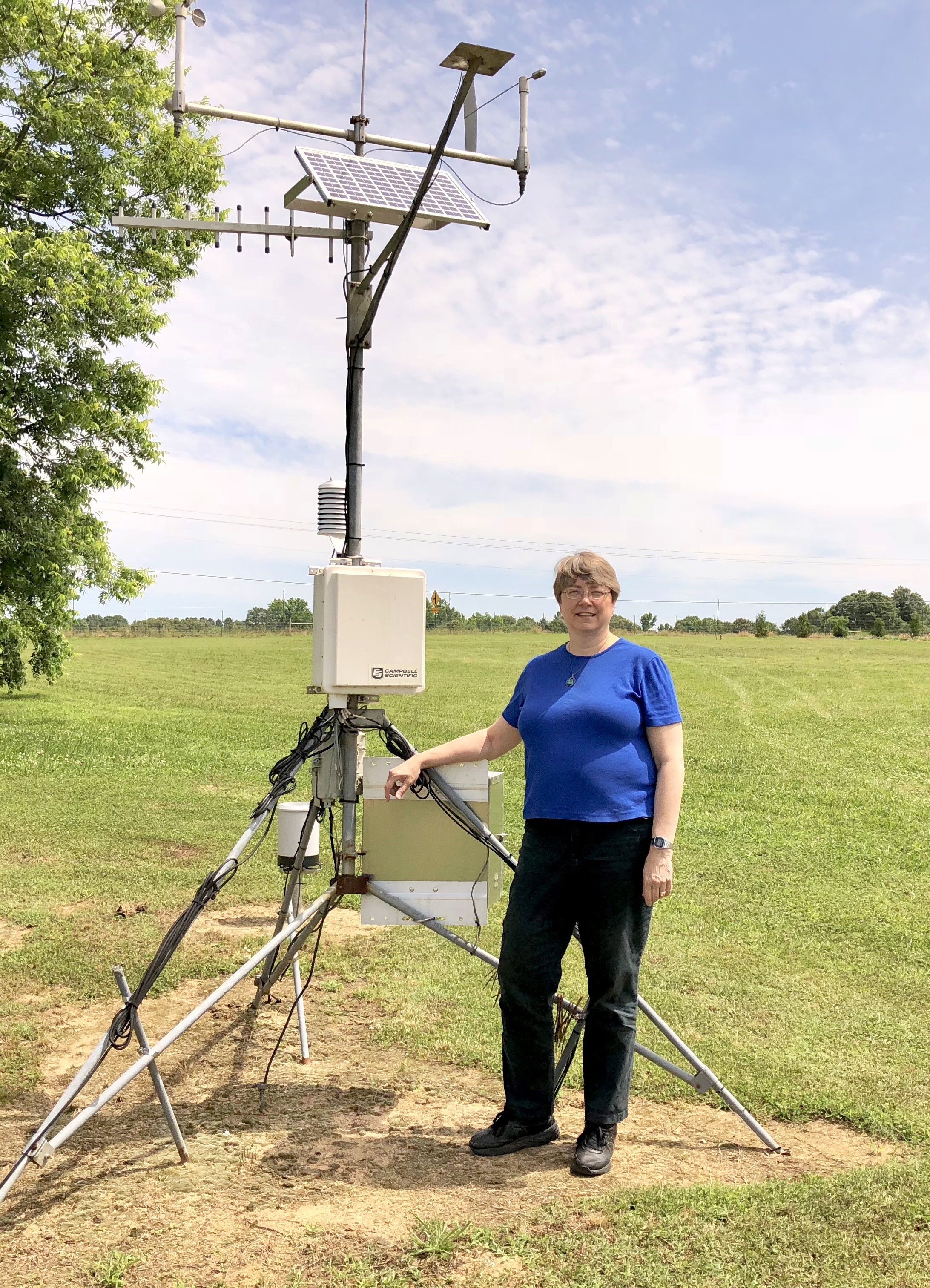 A woman with short hair and glasses, wearing a blue shirt, poses with a weather monitoring station in a field.