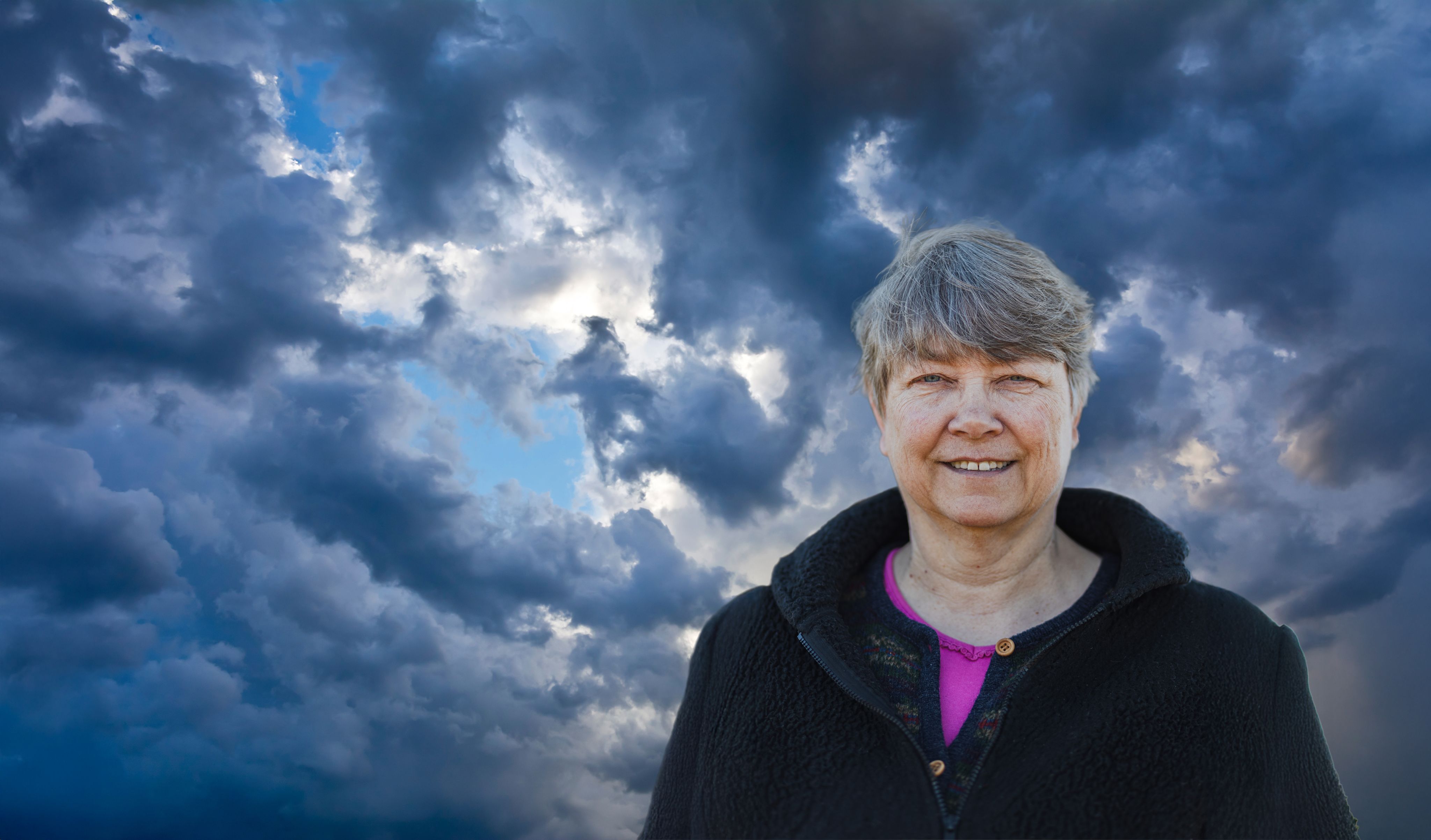 A smiling woman with short hair is farmed against a sky full of storm clouds in the background.