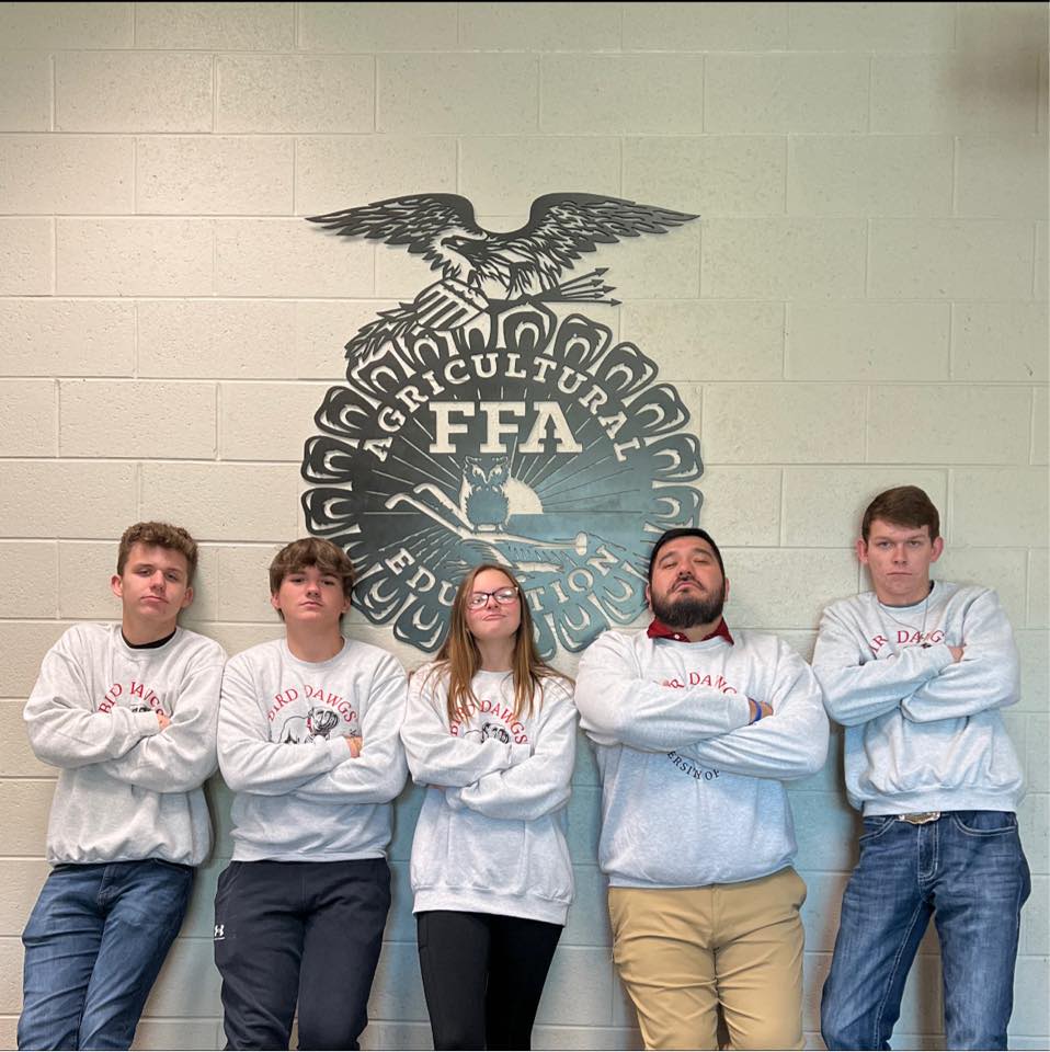 Pedraza and four of his students pose in UGA Bird Dawgs sweatshirts in front of the FFA agricultural education seal on the wall.
