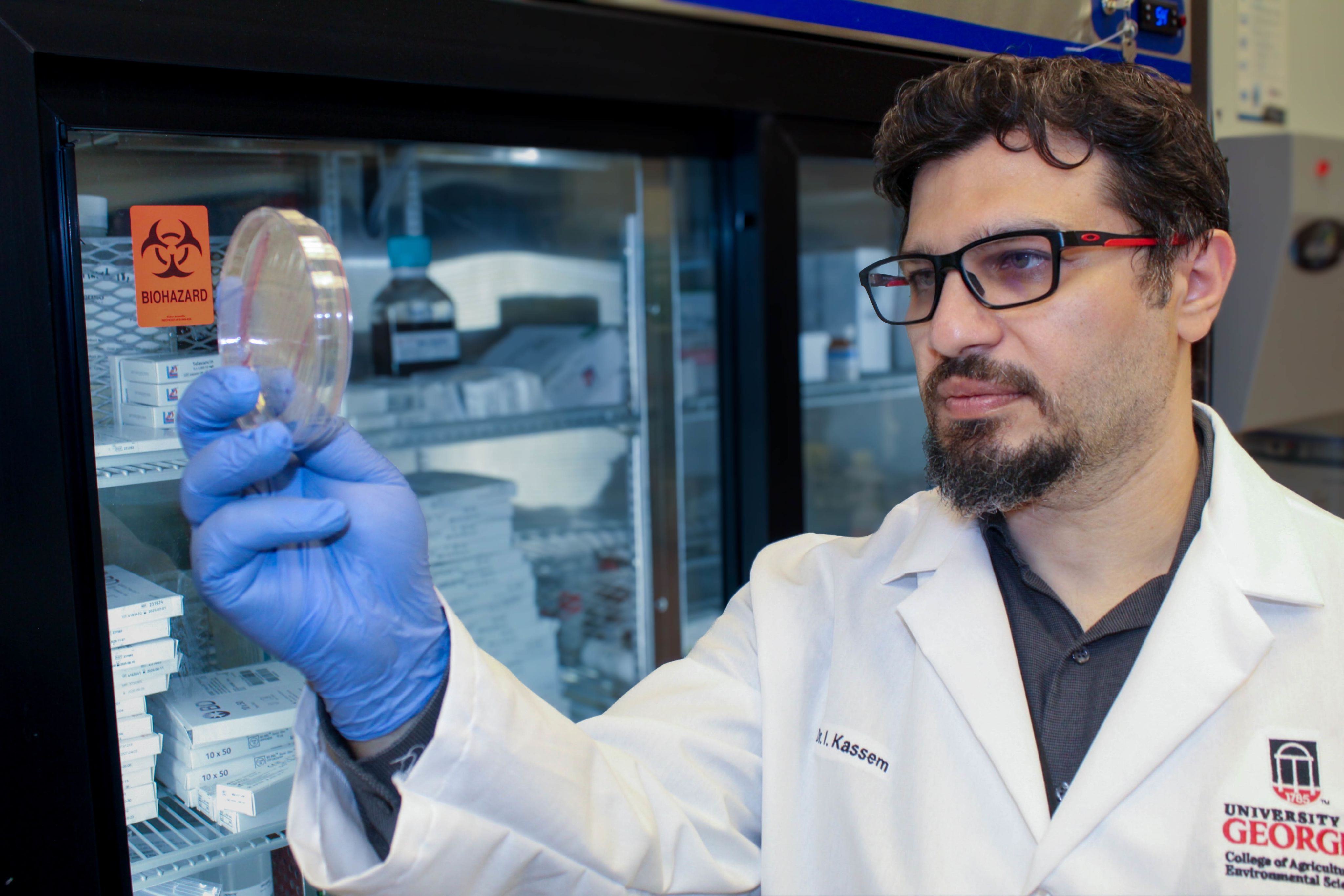 Issmat Kassem wears a white lab coat and blue gloves while holding up a petri dish in a lab setting.