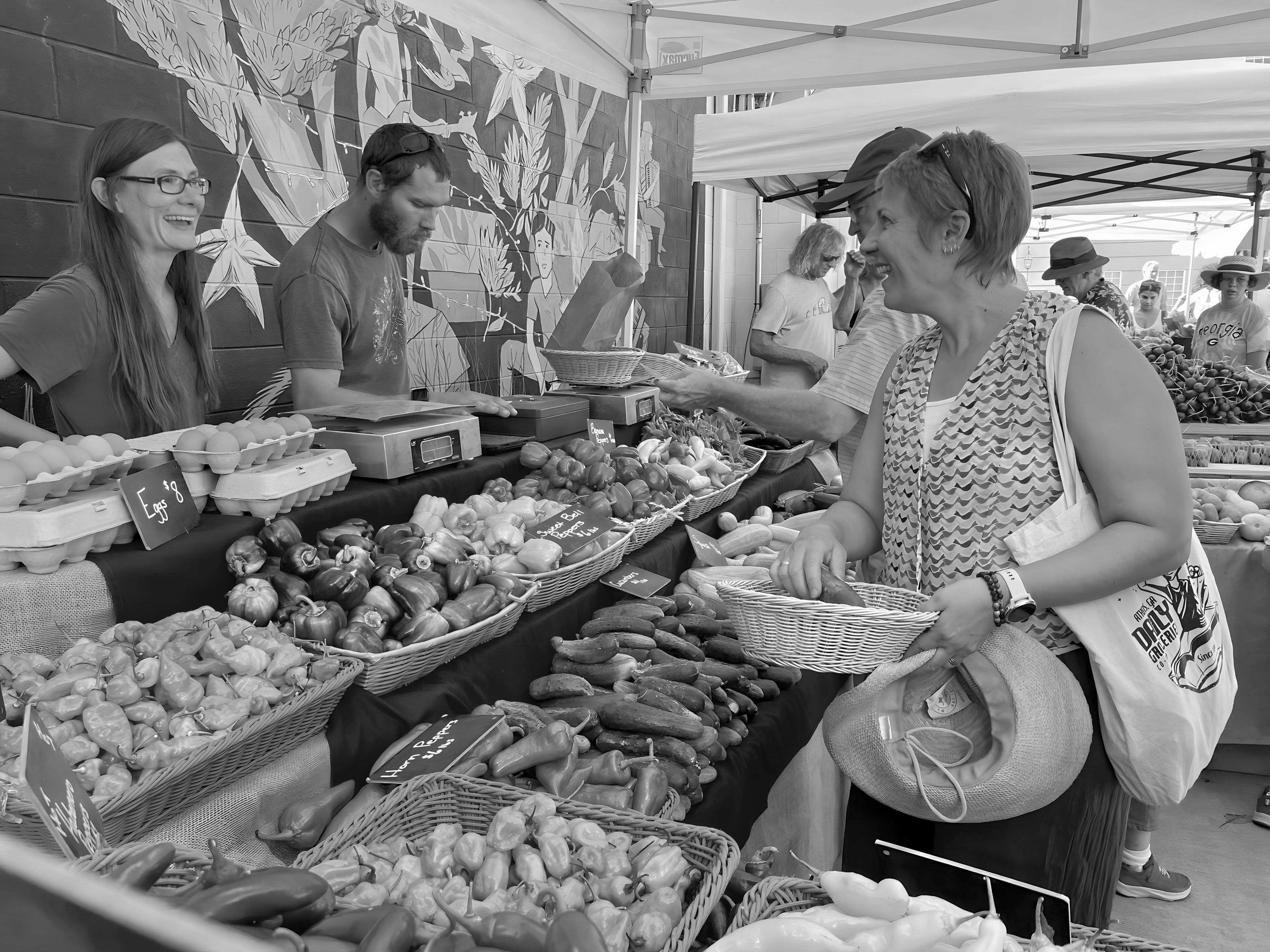 A black and white image of Thompson at a farmer's market. Thompson, on the right, is smiling as she talks to the market vendors who are selling various types of fresh produce. 