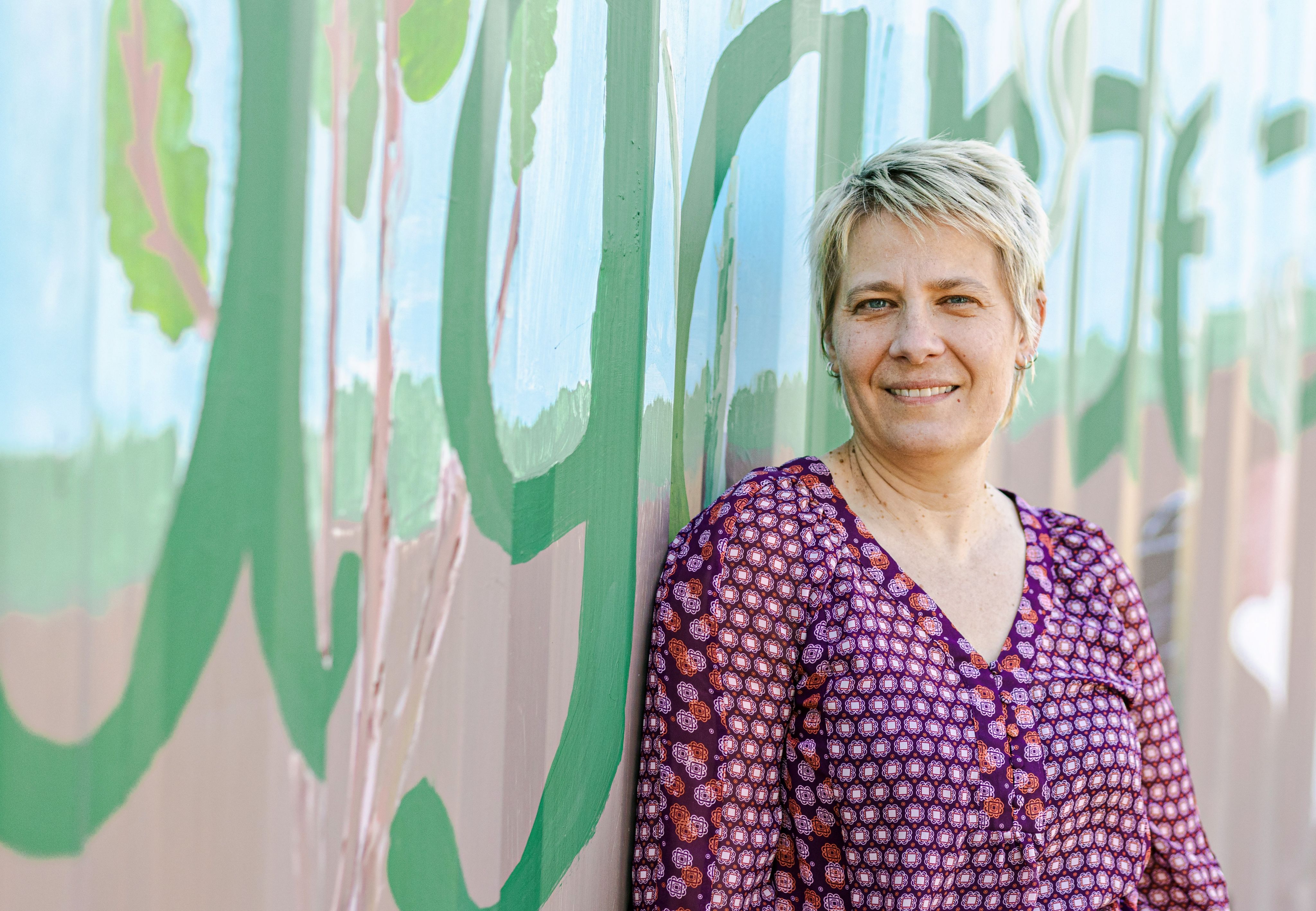 Jennifer Jo Thompson wears a purple top and stands against a blue wall with green writing painted on it. 