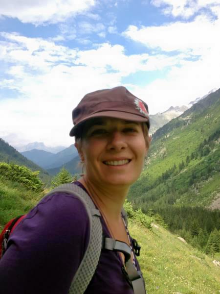Thompson is wearing a hat and backpack with green mountainous terrain in the background. The blue sky above has several white clouds. 