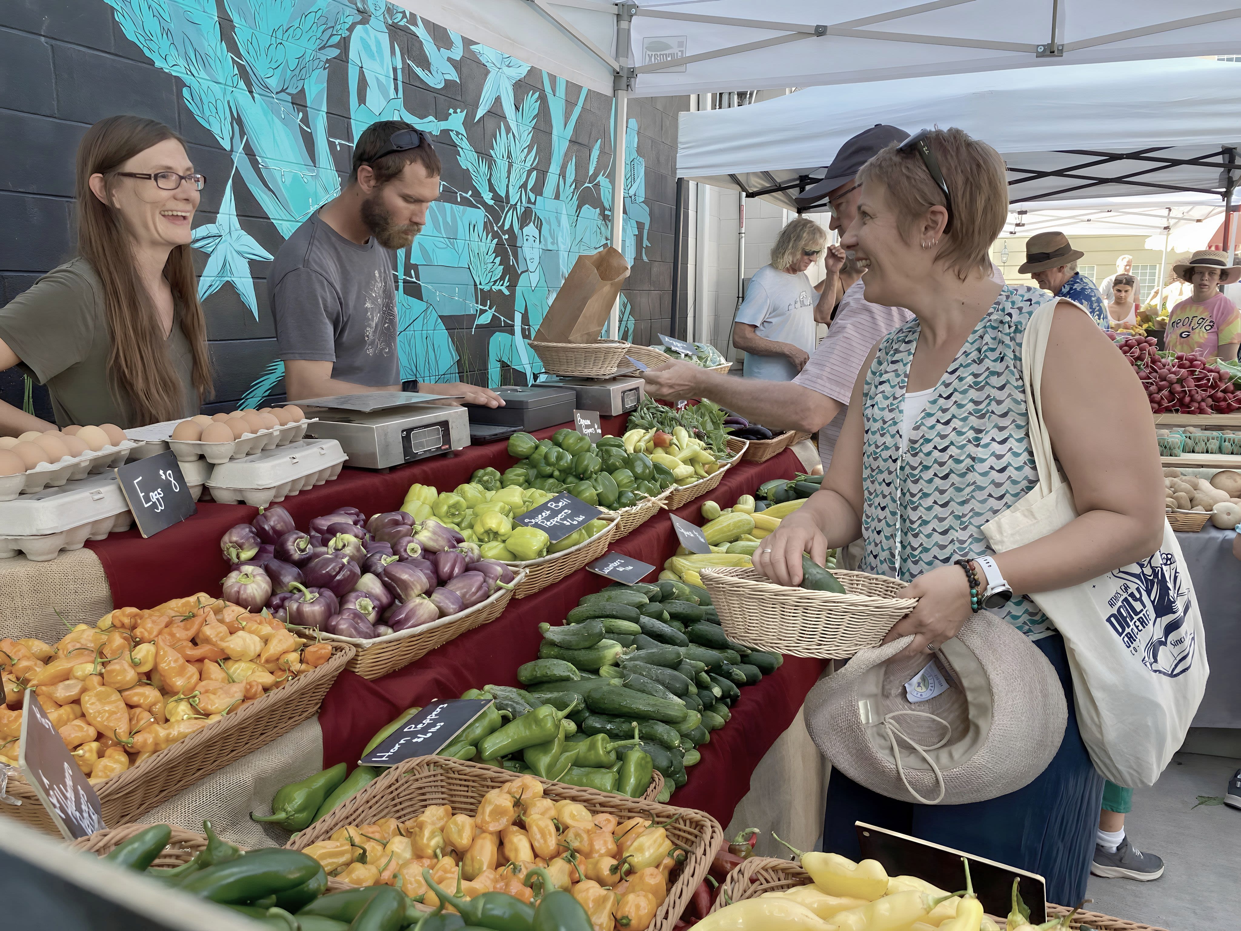 The black and white image begins to become saturated with color, which shows Thompson at a farmer's market. Thompson, on the right, is smiling as she talks to the market vendors who are selling various types of fresh produce. 