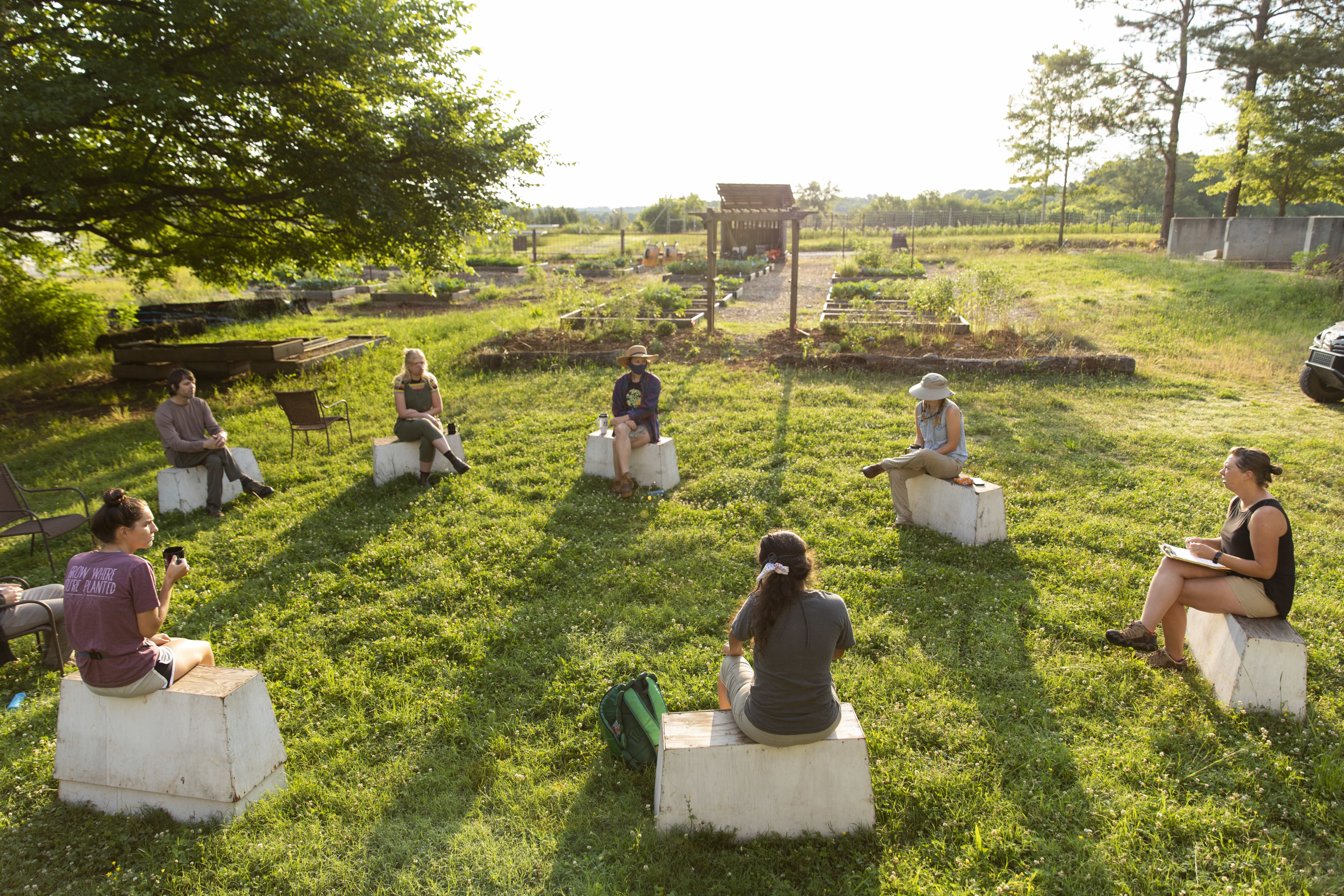 Farm manager JoHannah Biang, right, talks with staff as they sit together on seats that are six feet apart during a morning meeting at the UGArden.