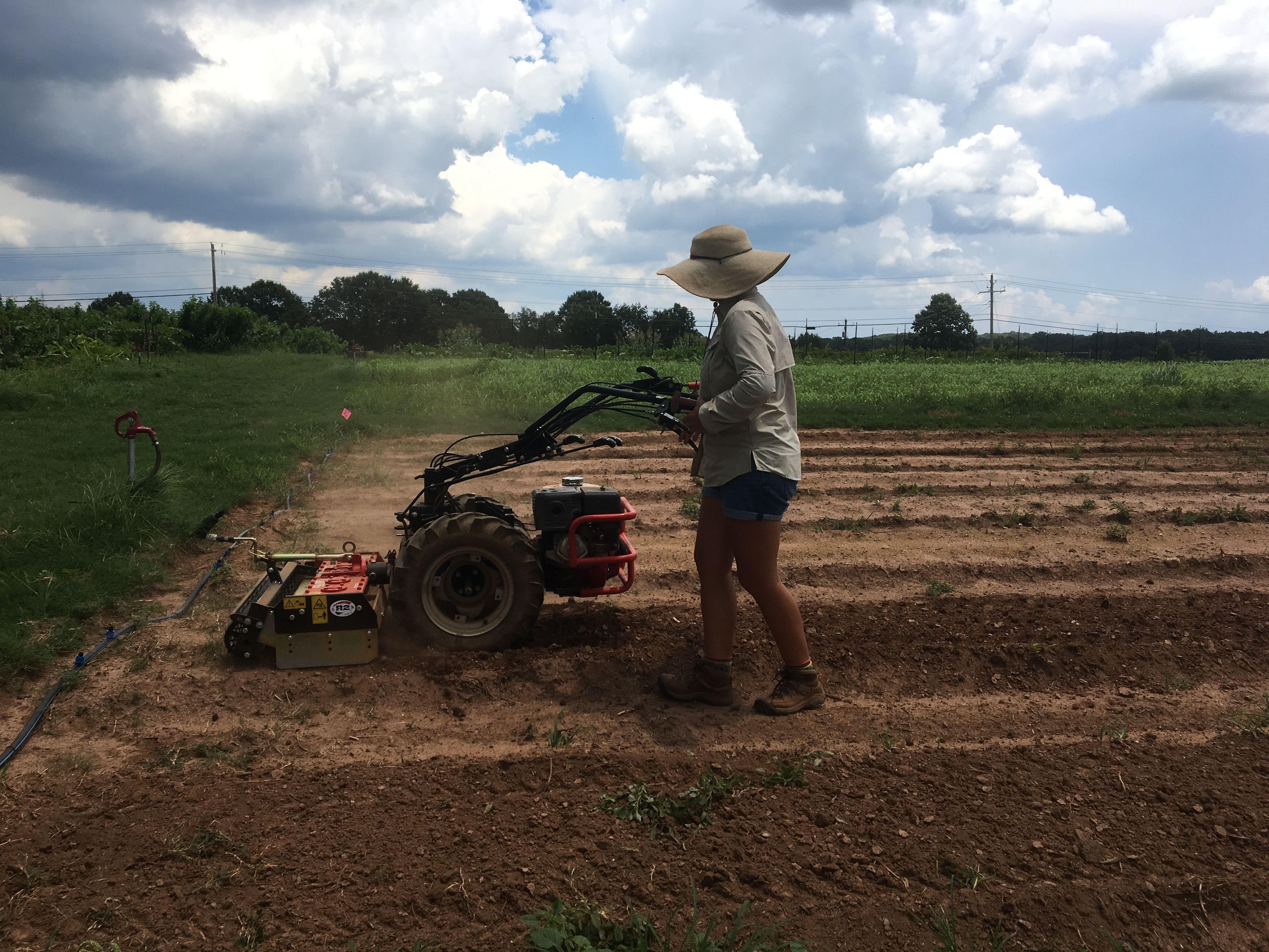 JoHannah Biang uses a motorized tiller in a garden plot, while wearing a sun hat, long-sleeved shirt, and shorts.