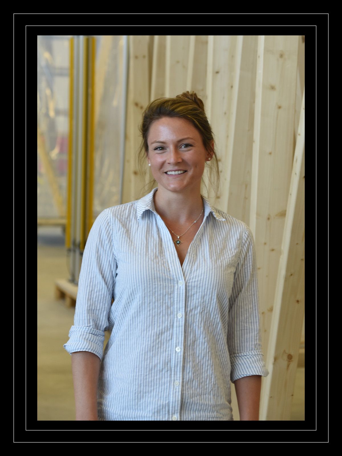 Kaitlin McNally smiles in front of standing lumber wearing a button-up shirt