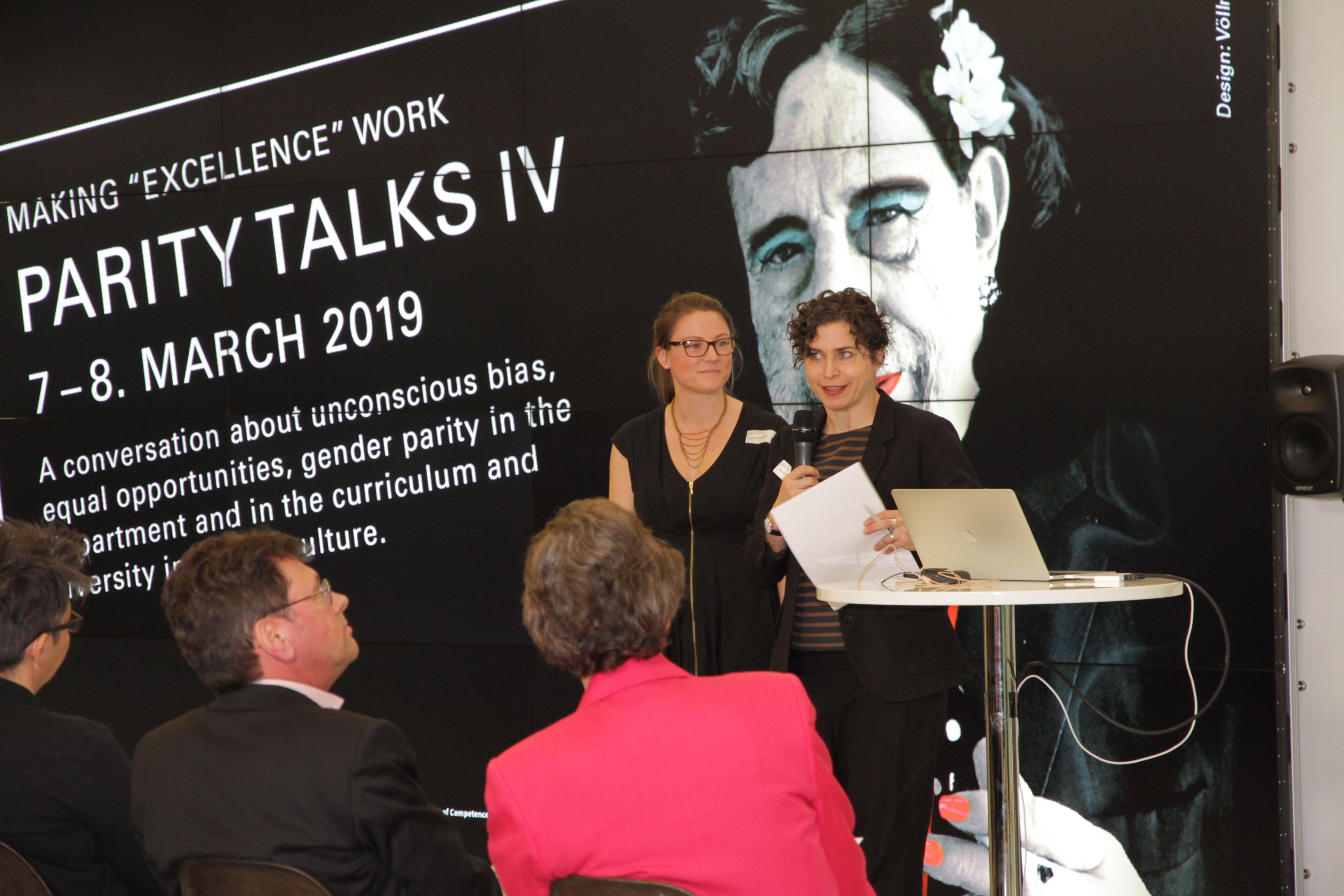 Kaitlin McNally stands at a modern lectern with a speaker at the Parity Talks IV event at ETH Zurich