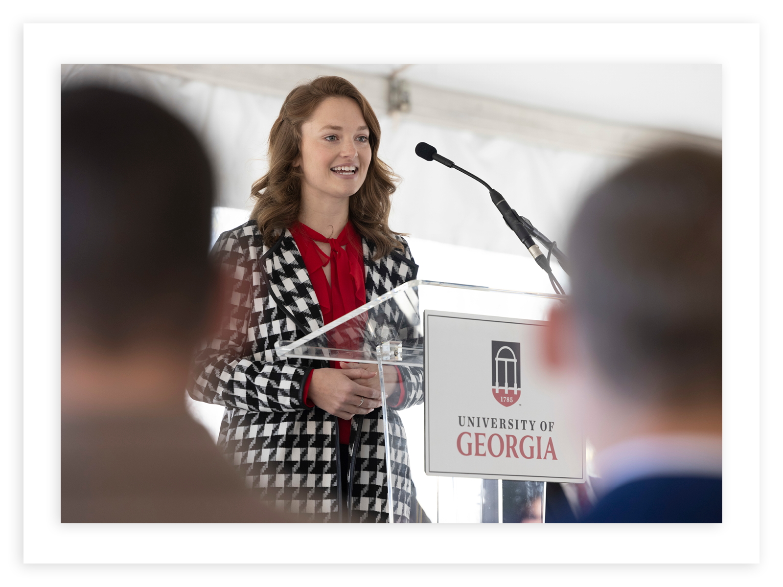 UGA student Kylie Bruce stands before a clear lectern with a University of Georgia logo on the front. There are two out of focus heads in the foreground and Kylie is wearing a red top with a houndstooth coat