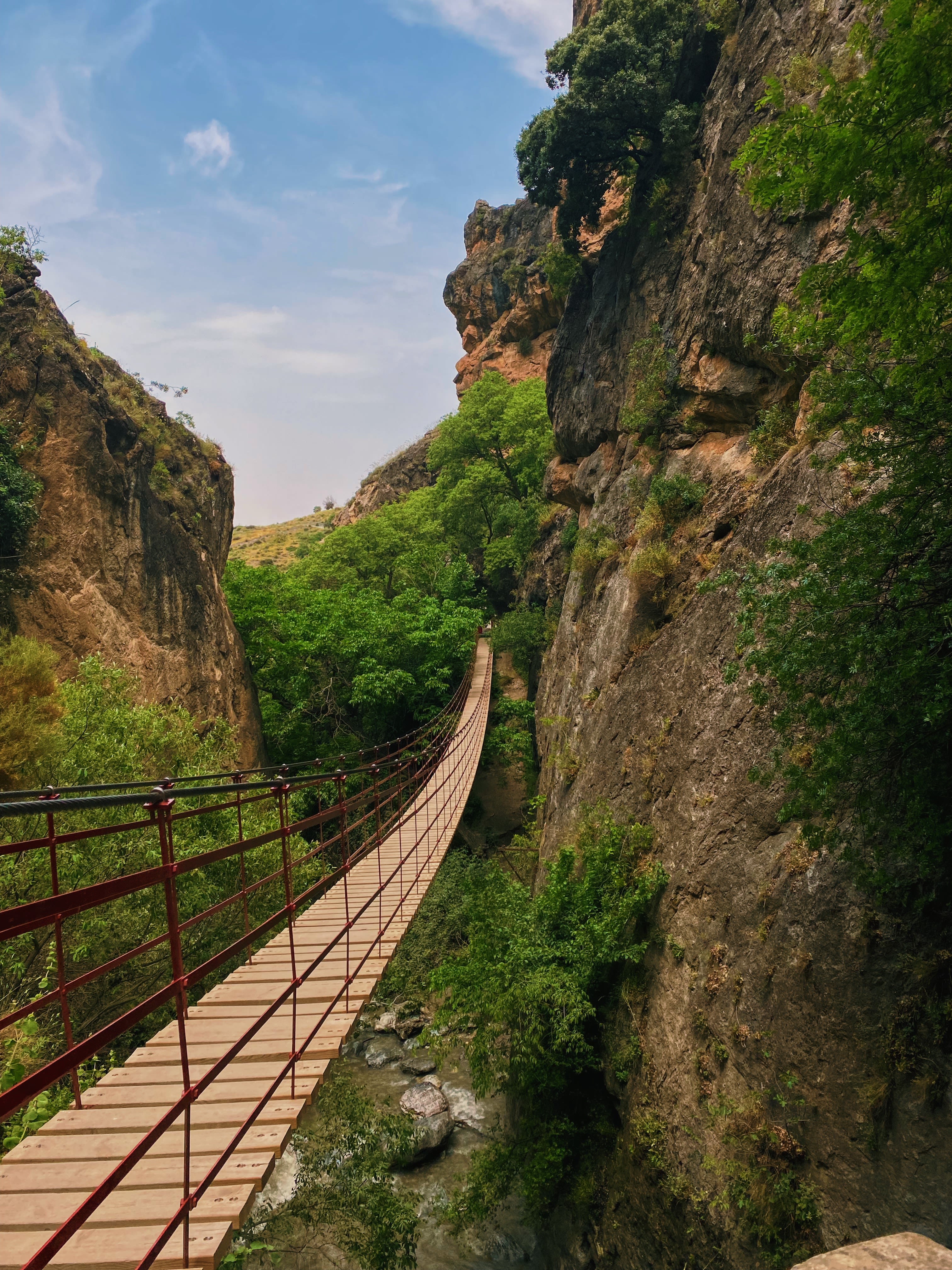 A foot bridge in Spain goes between two rock cliffs in Spain, with leafy trees visible around the bridge.