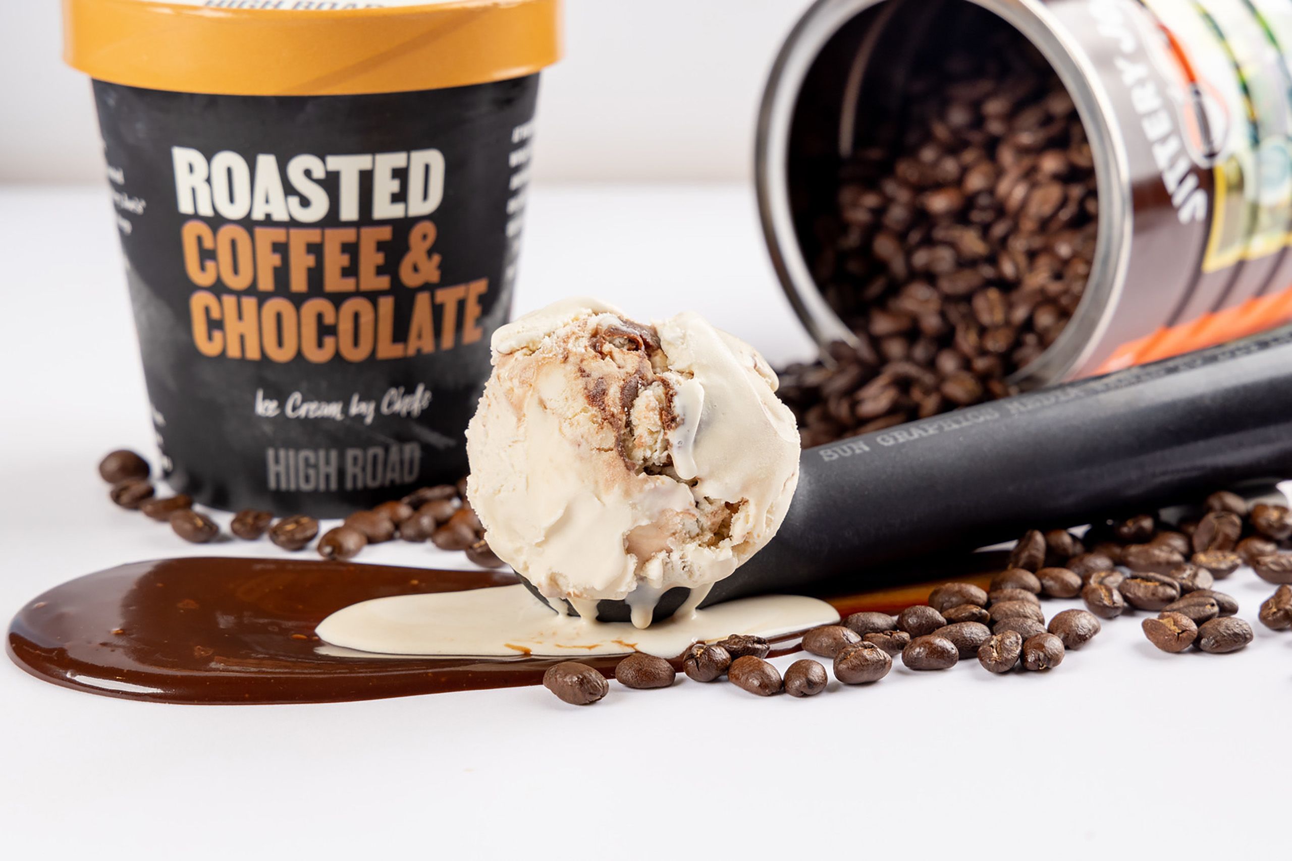 A scoop of roasted coffee and chocolate ice cream melts into a pool of chocolate syrup, surrounded by a scatter of coffee beans.