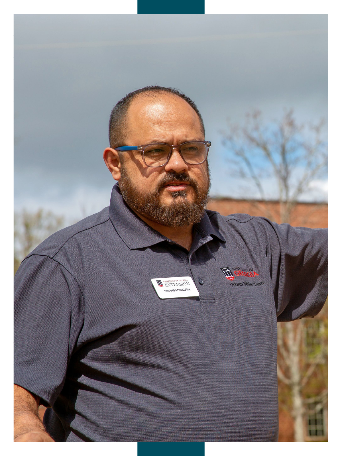 Rolando Orellana stands in front of brick buildings with his arm extended as he talks about the irrigation demonstration plots on Griffin Campus.