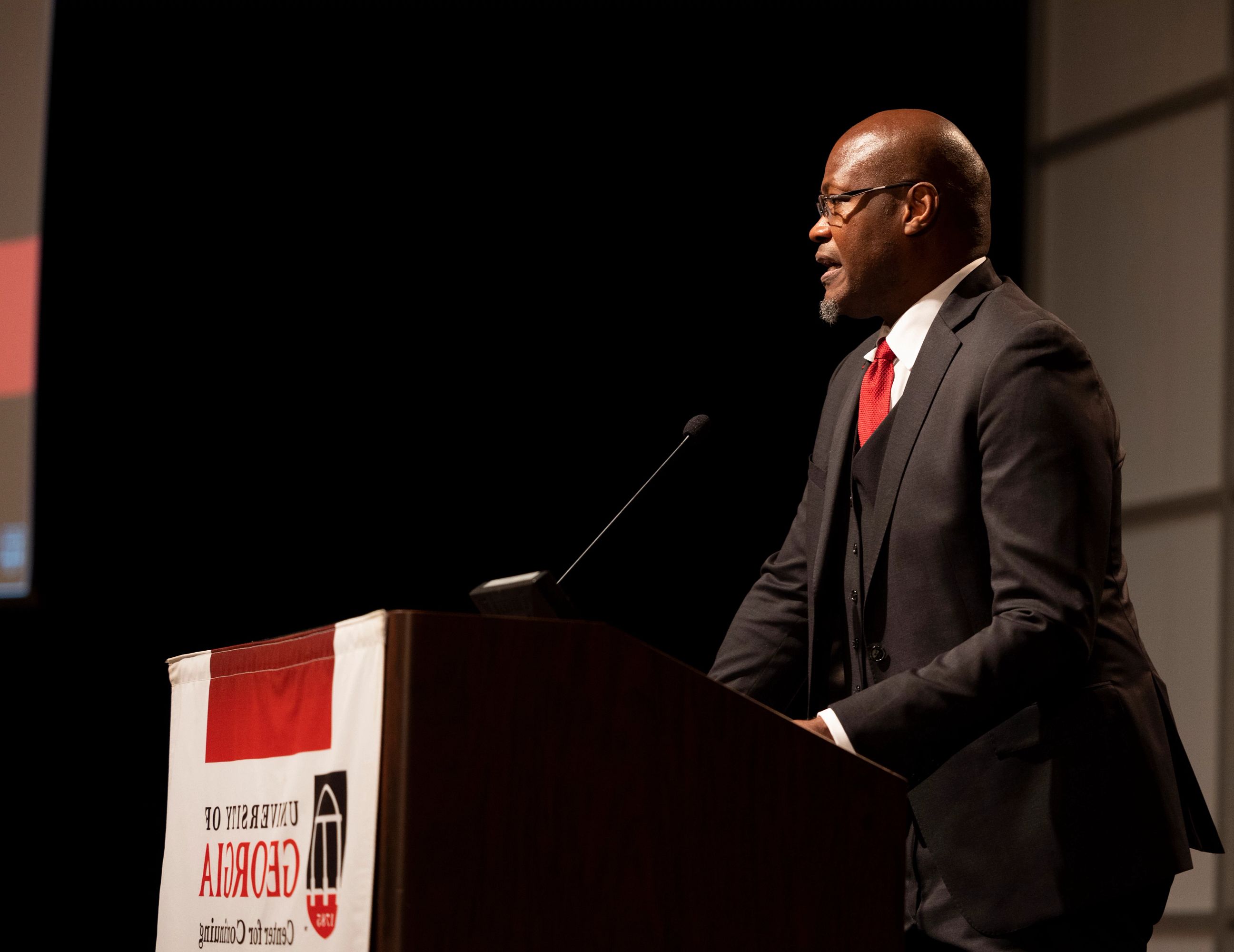 Walcott speaks at the 23rd Mary Frances Early Lecture in 2023. He stands in front of the crowd in a darkened room. He stands at the podium to speak, wearing a black suit, red tie, white collared shirt and glasses.