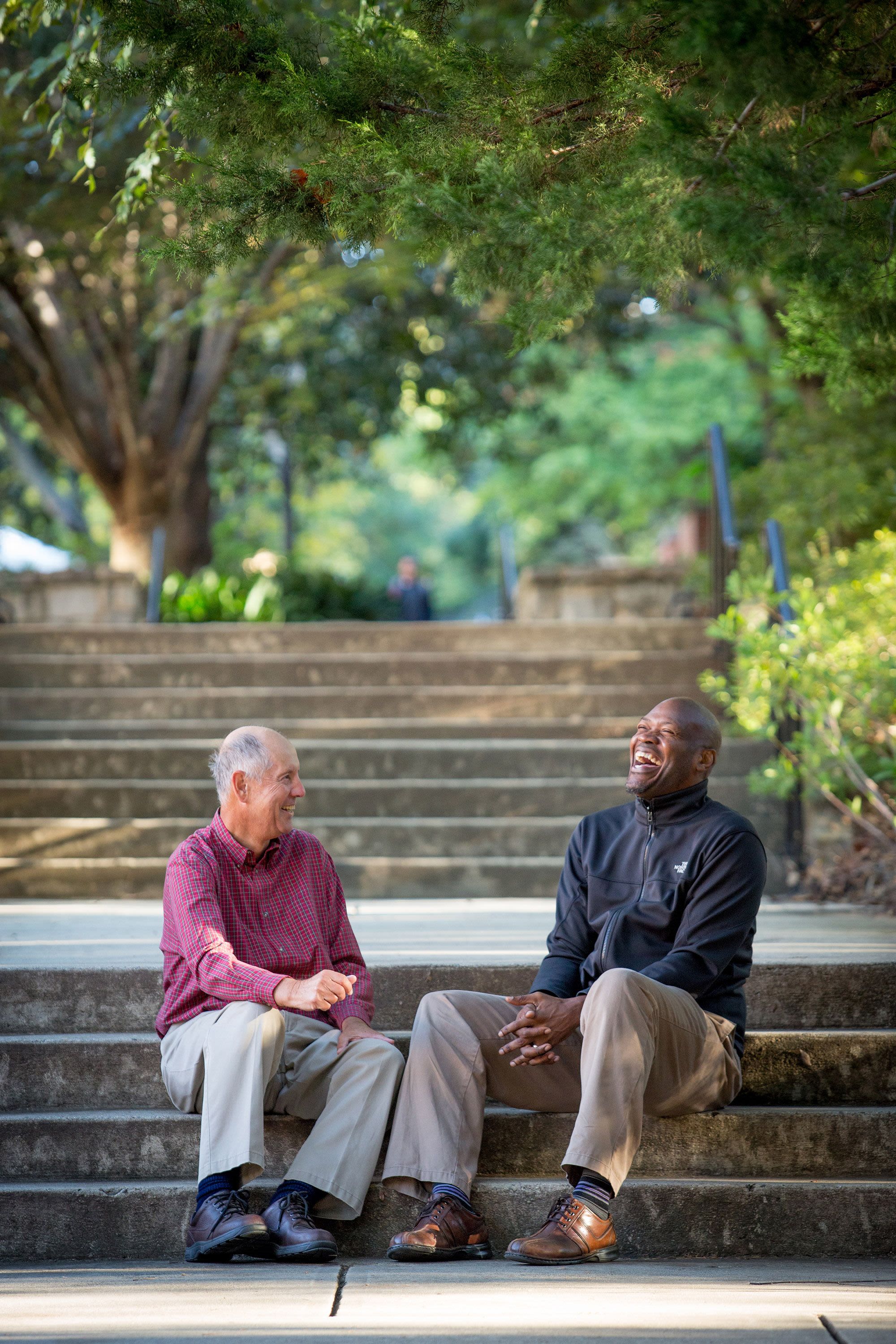 Walcott sits on stairs with a colleague on a sunny day. Walcott is laughing and the other man is smiling.