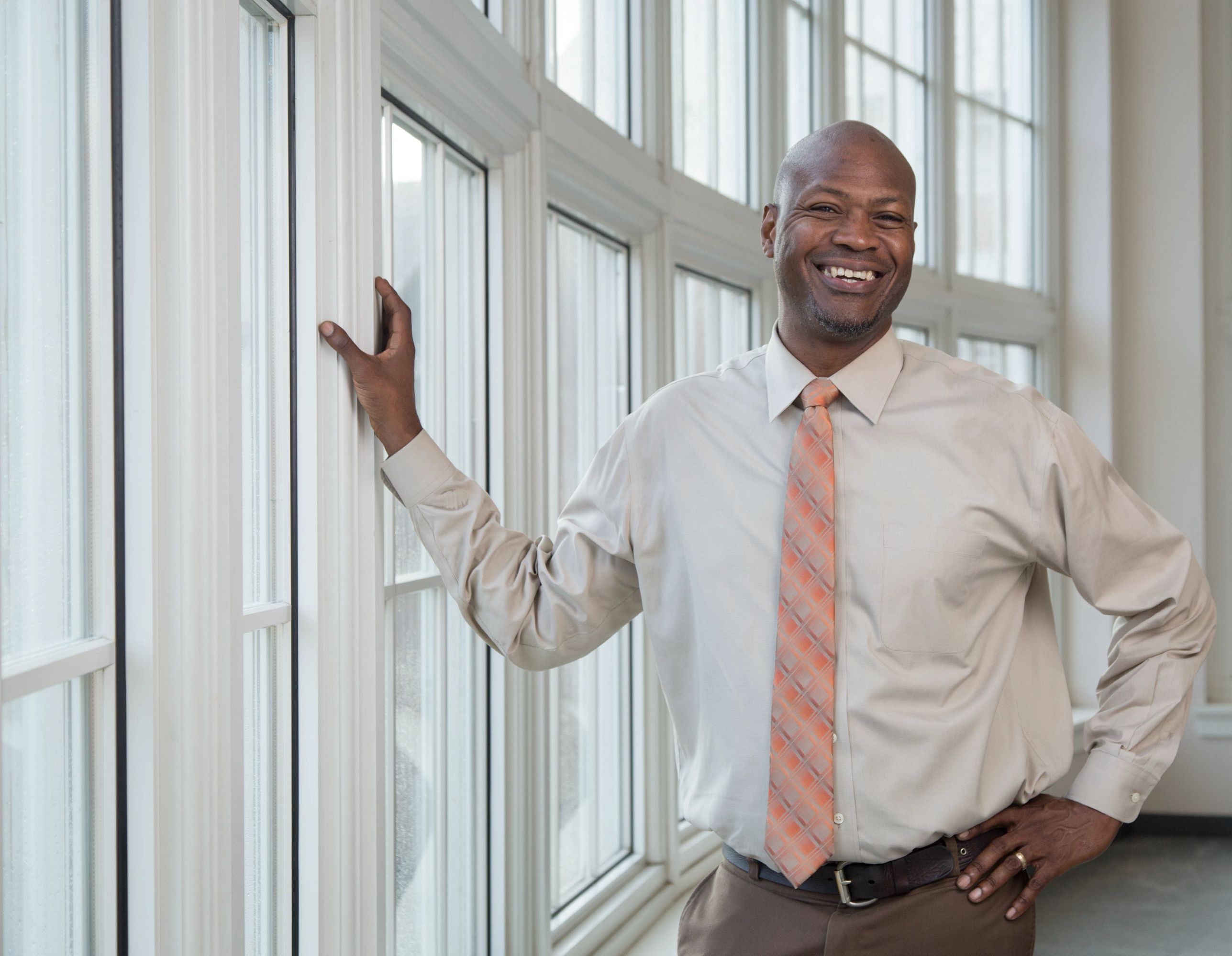 Walcott stands in a room with windows. He is smiling and looking at the camera. He wears a white shirt with an orange tie.