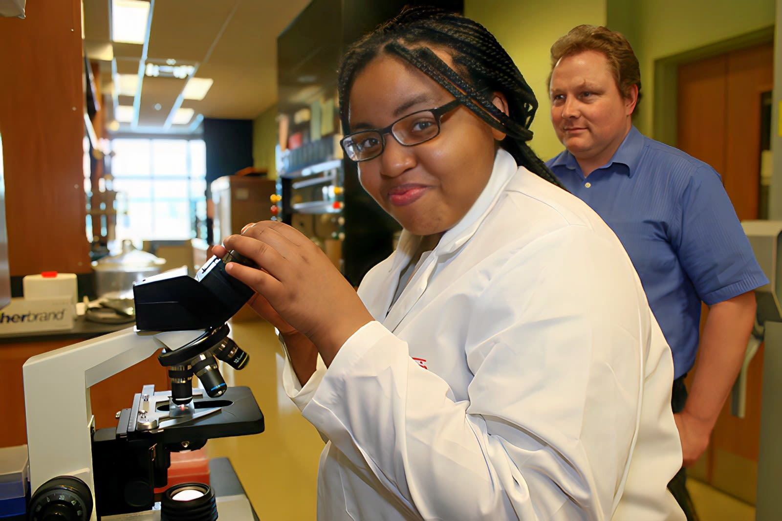 A student smiles as she looks up from the microscope. She's wearing a lab coat and glasses. A man with a blue shirt stands behind her looking at the microscope.