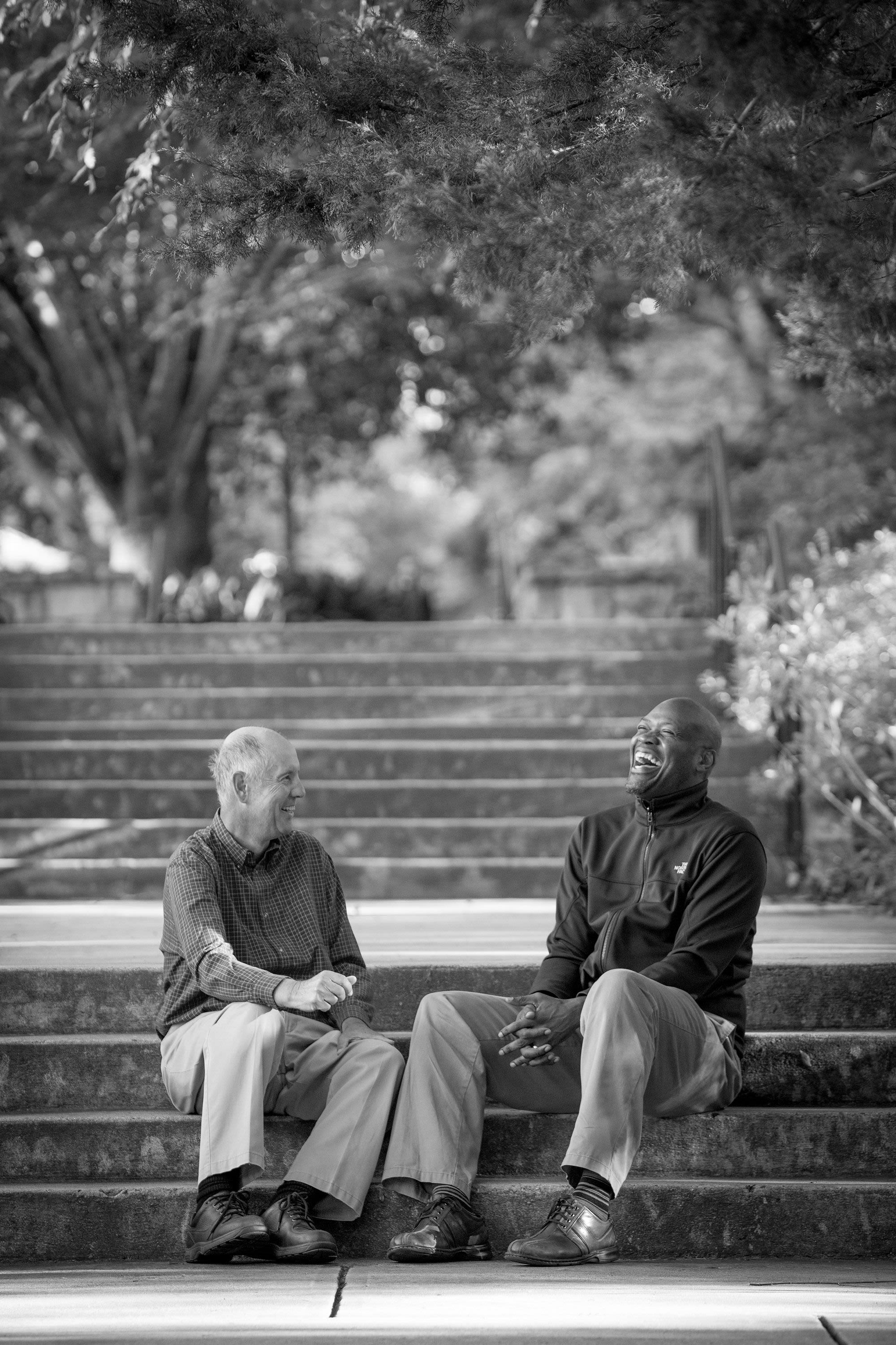 Walcott sits on stairs with a colleague on a sunny day. Walcott is laughing and the other man is smiling.
