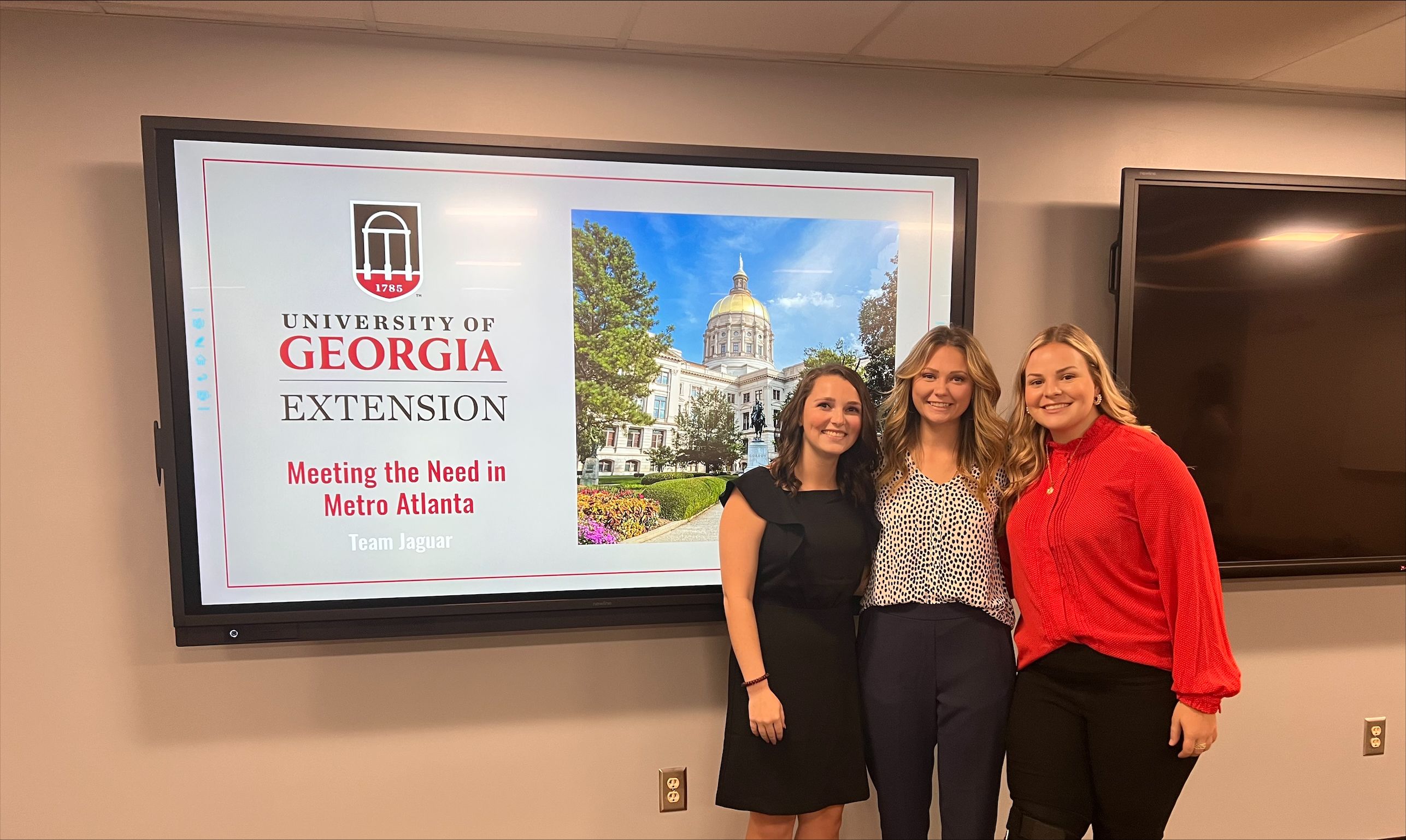 Three students stand in front a presentation screen that reads "University of Georgia Extension, Meeting the Need in Metro Atlanta, Team Jaguar.