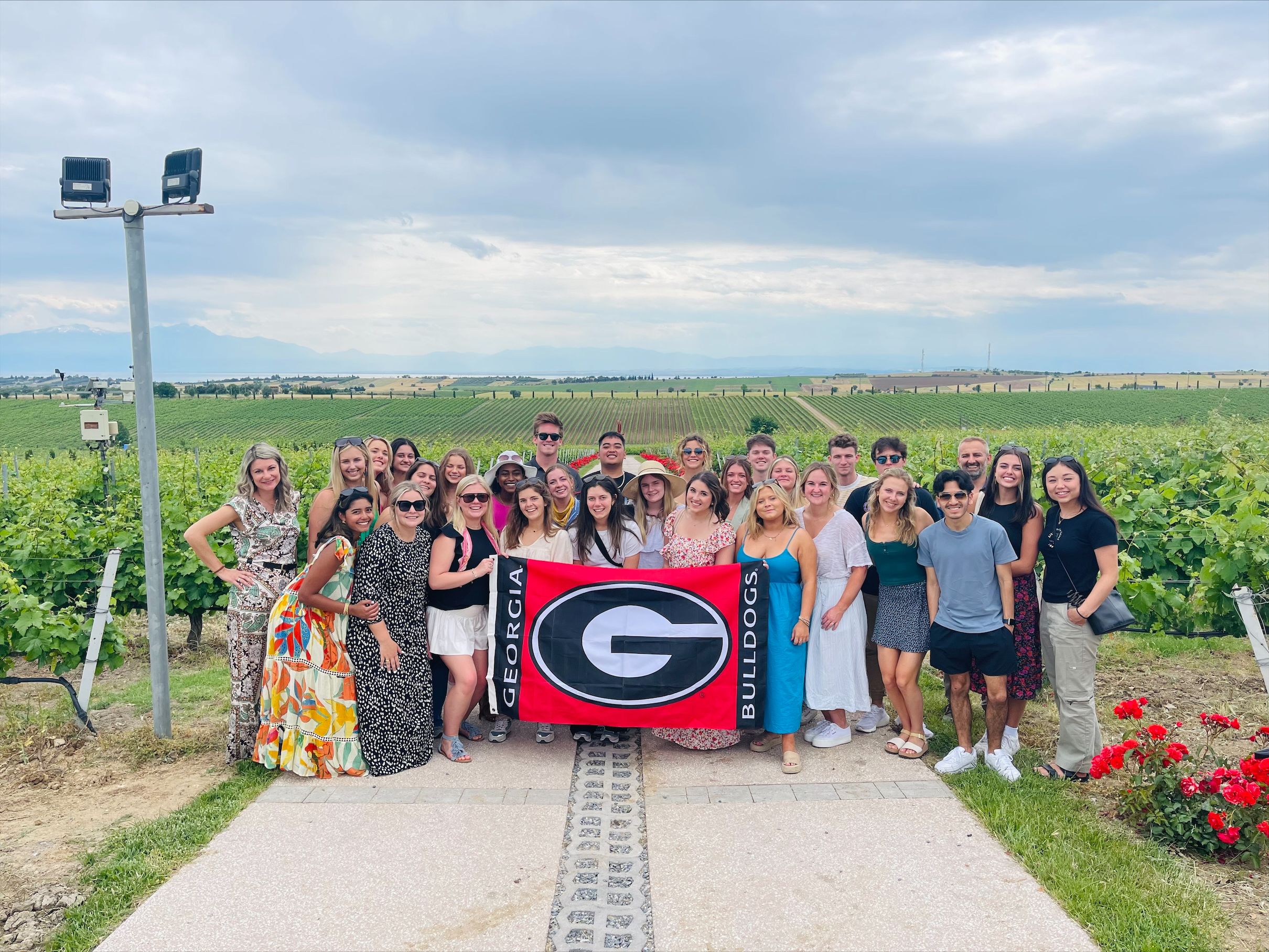 The study abroad group holds a Georgia Bulldogs flag with UGA's Super G in the middle. Behind the group of students is a vineyard on a partly cloudy day.