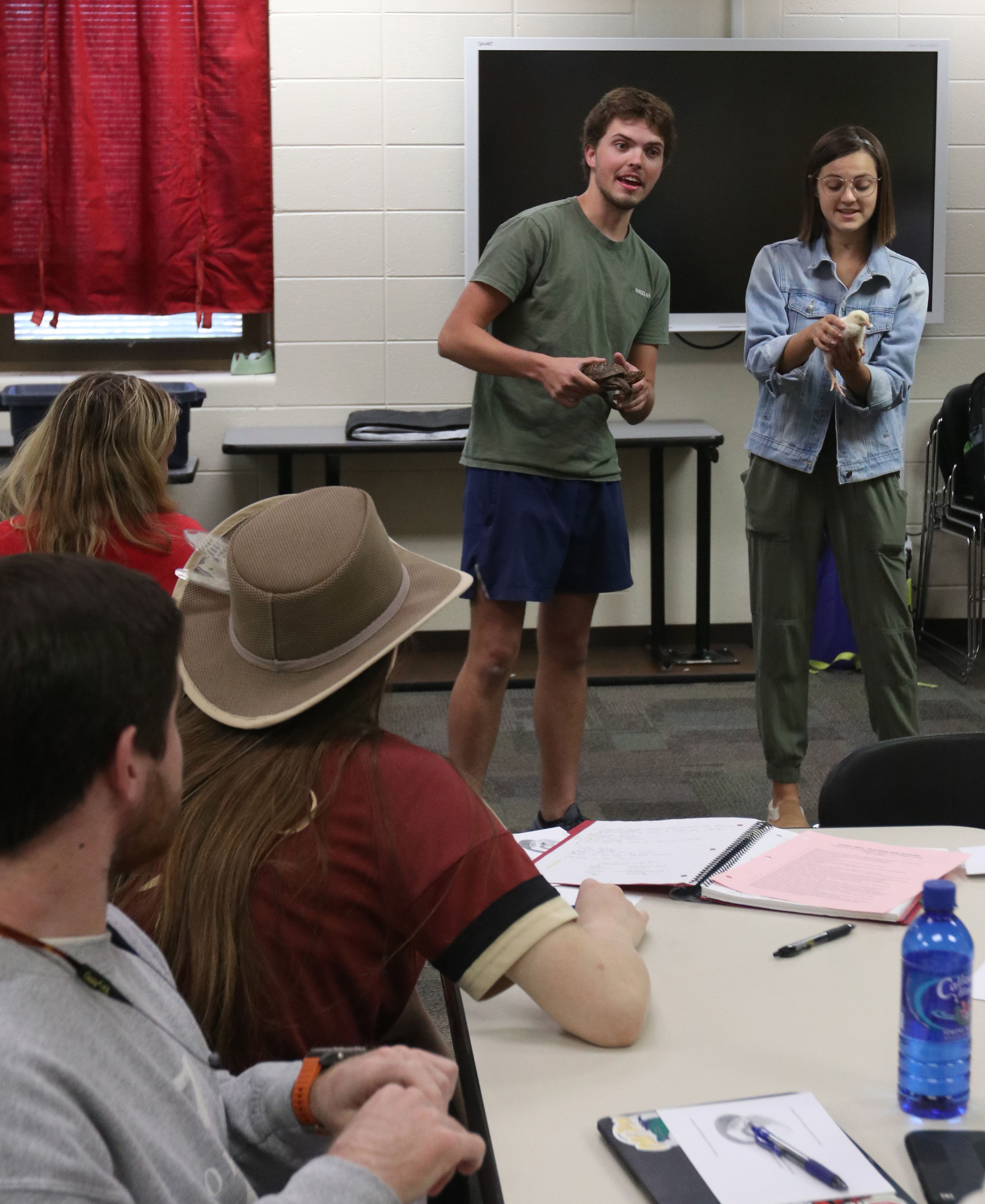 Two students stand in front of the class holding a turtle and a chick while demonstrating a lesson
