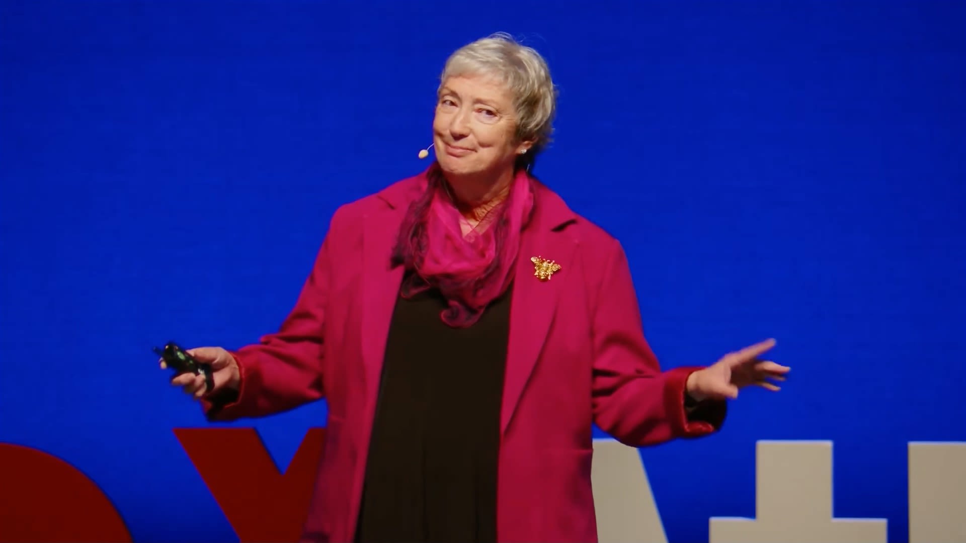Kris Braman smiles with a blue screen behind her. She's wearing a red coat and gold bee pendant.