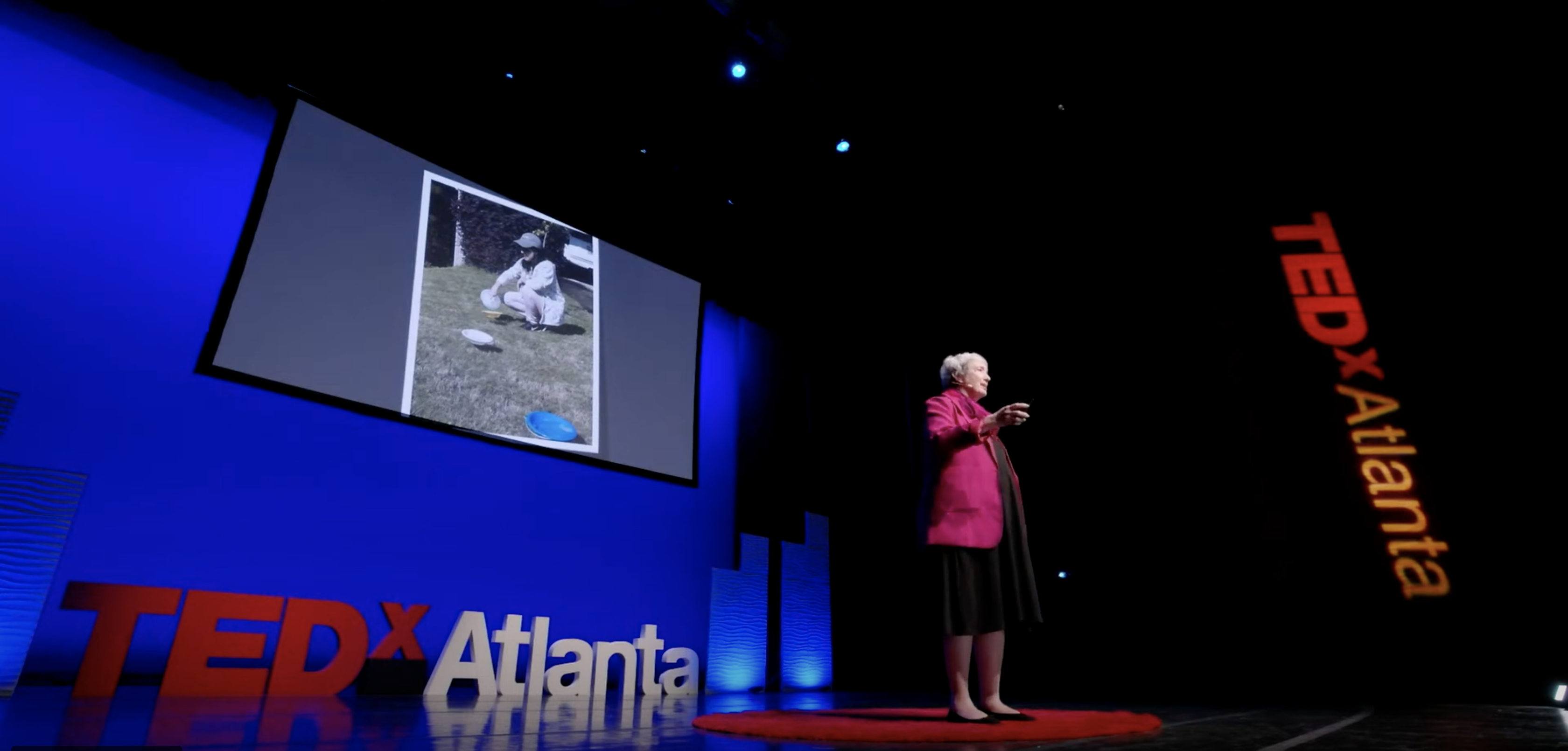 Kris Braman stands on stage at the TEDxAtlanta event, photo is taken from below stage looking up at Braman, who is wearing a red coat.