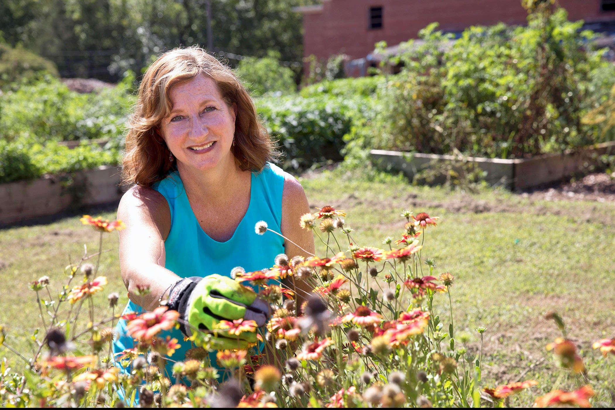 Wearing a bright blue shirt, Becky Griffin smiles as she reaches for a group of orange and yellow flowers. A couple raised garden beds can be seen in the background.