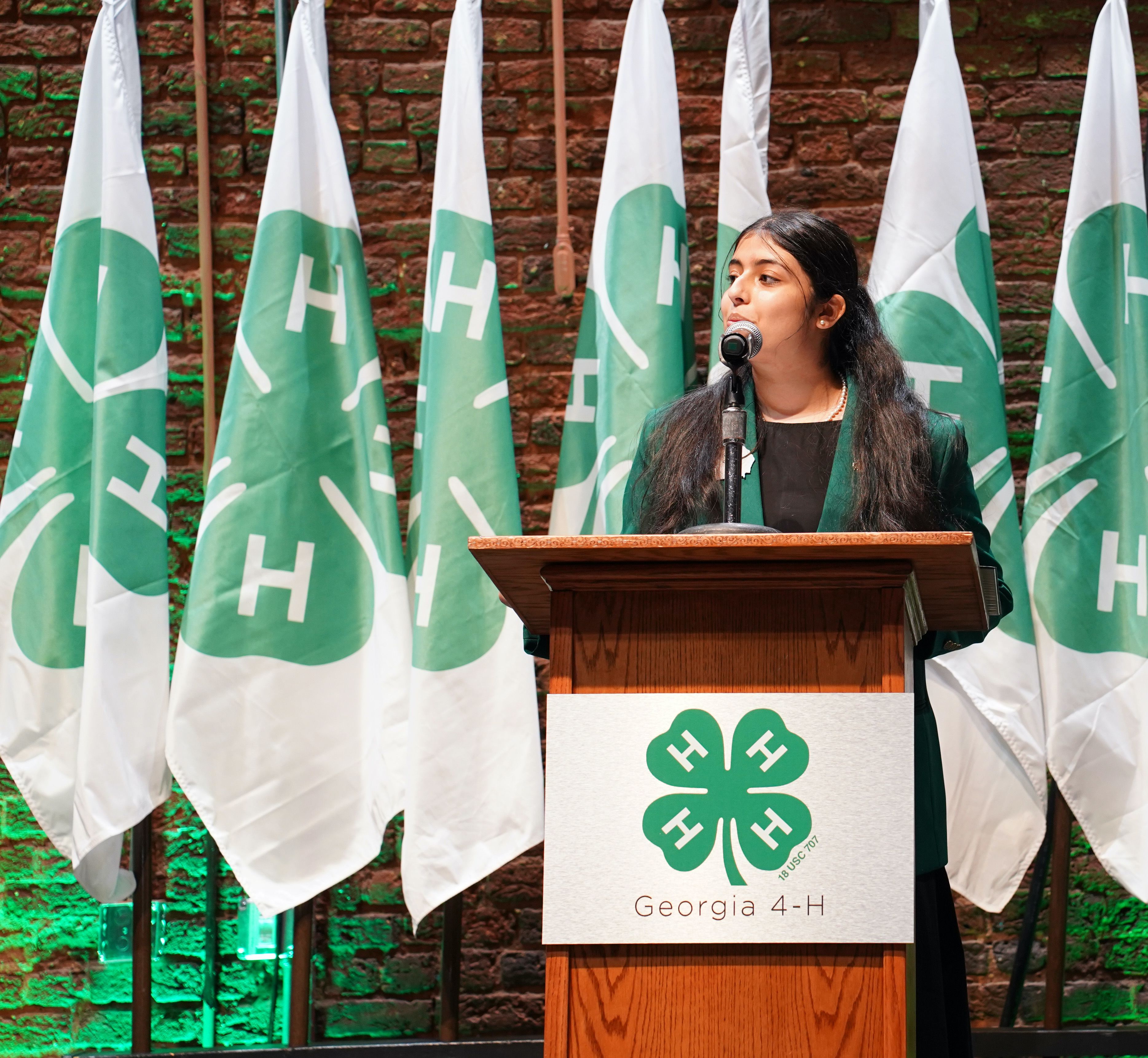 Gunjal speaks at a lectern in front of several flags bearing the 4-H emblem