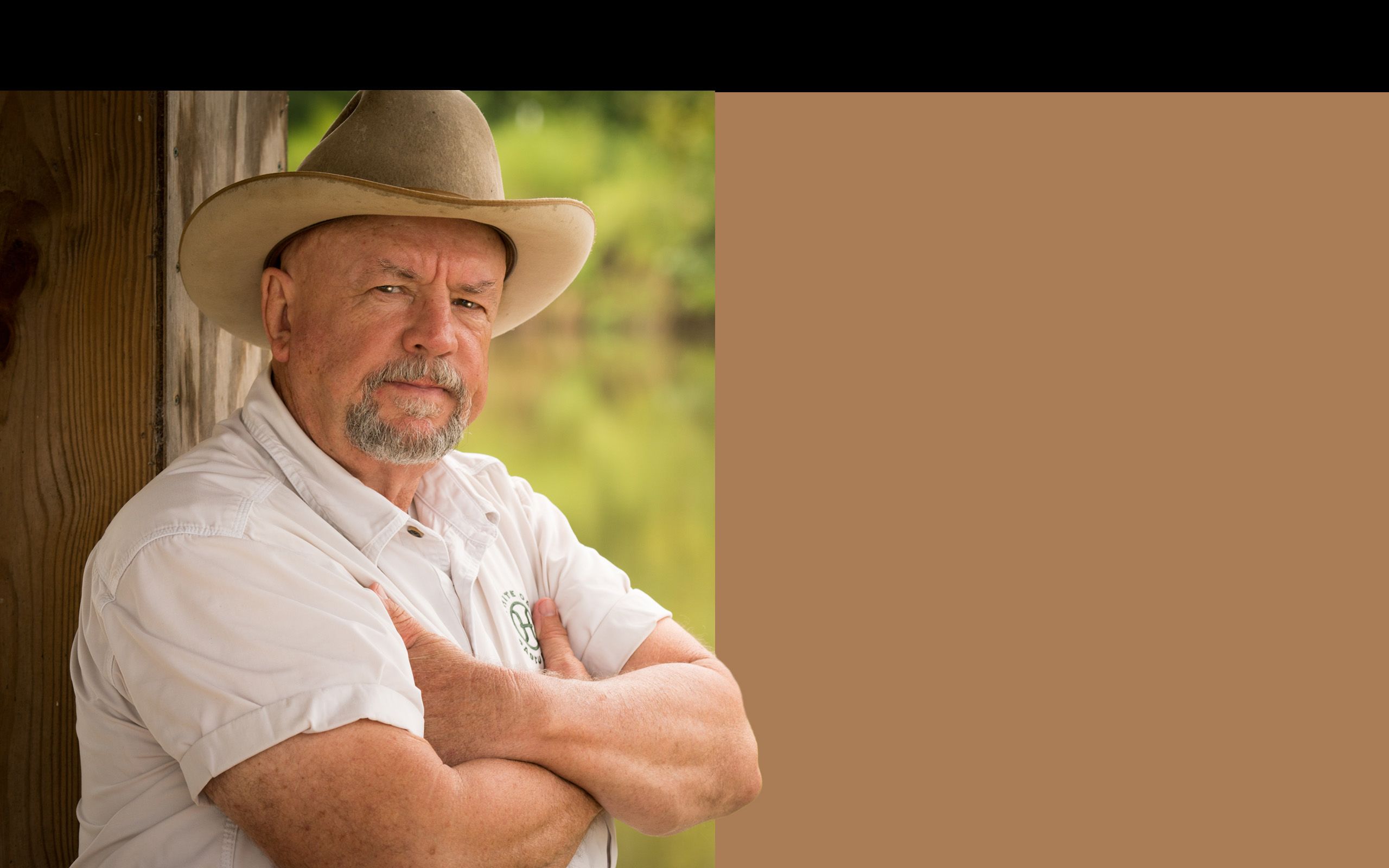 Portrait of Will Harris standing in a doorway with his arms folded across his chest wearing a white button up shirt and a well-worn tan cowboy hat